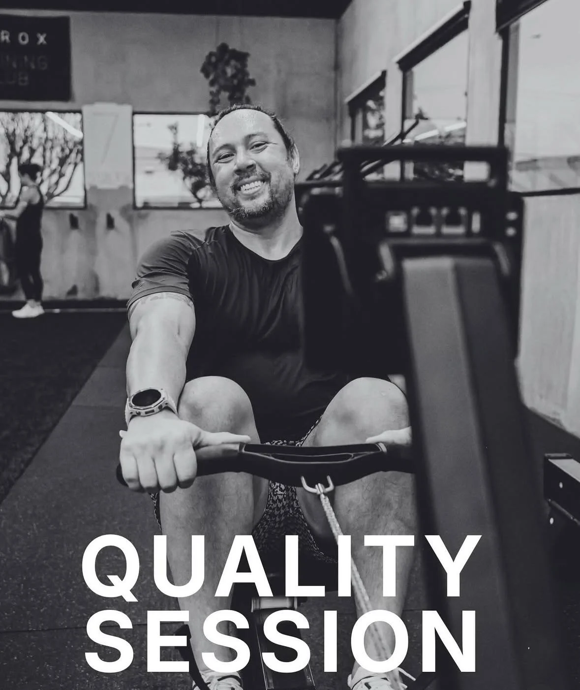 Man smiling while seated on a stationary bike in a gym during a fitness session, with a woman exercising in the background and the words 'QUALITY SESSION' overlaid.