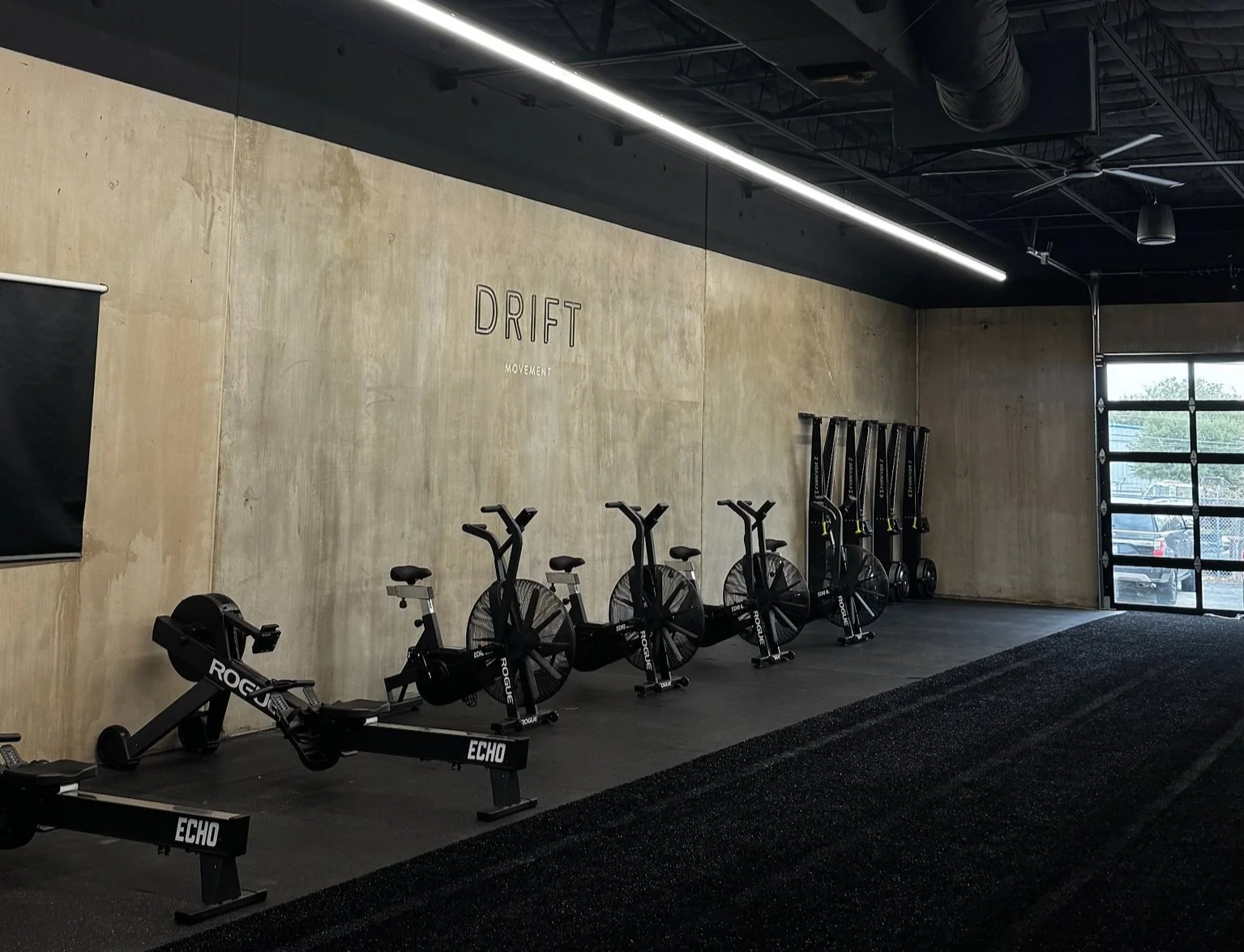 An empty gym with stationary bikes lined up against a concrete wall, with a black rubber floor and a large window at the end of the room.