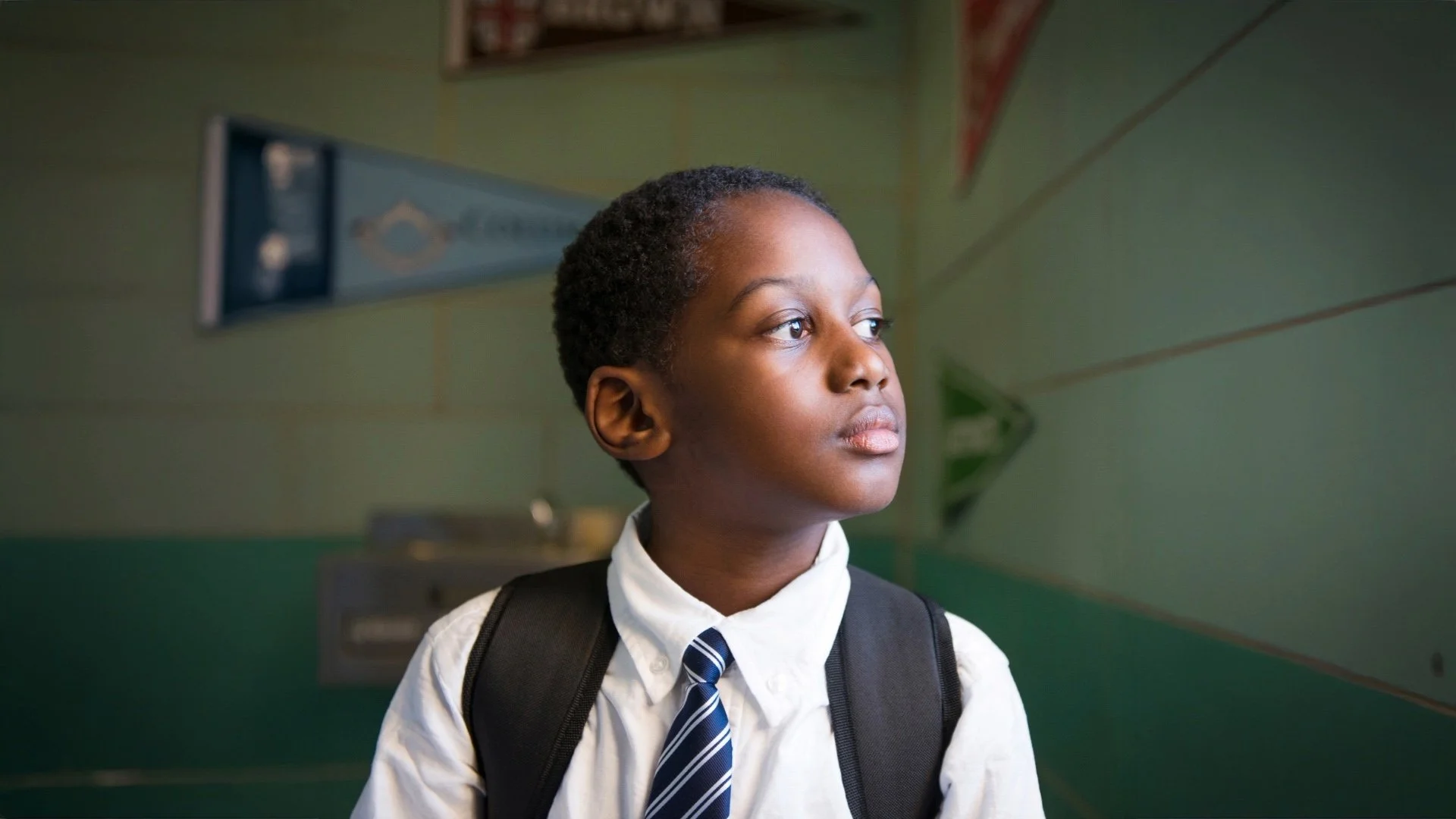 A young boy with dark skin and short curly hair wearing a white shirt, a striped tie, and a backpack, standing in a school hallway with green walls and looking to the side.