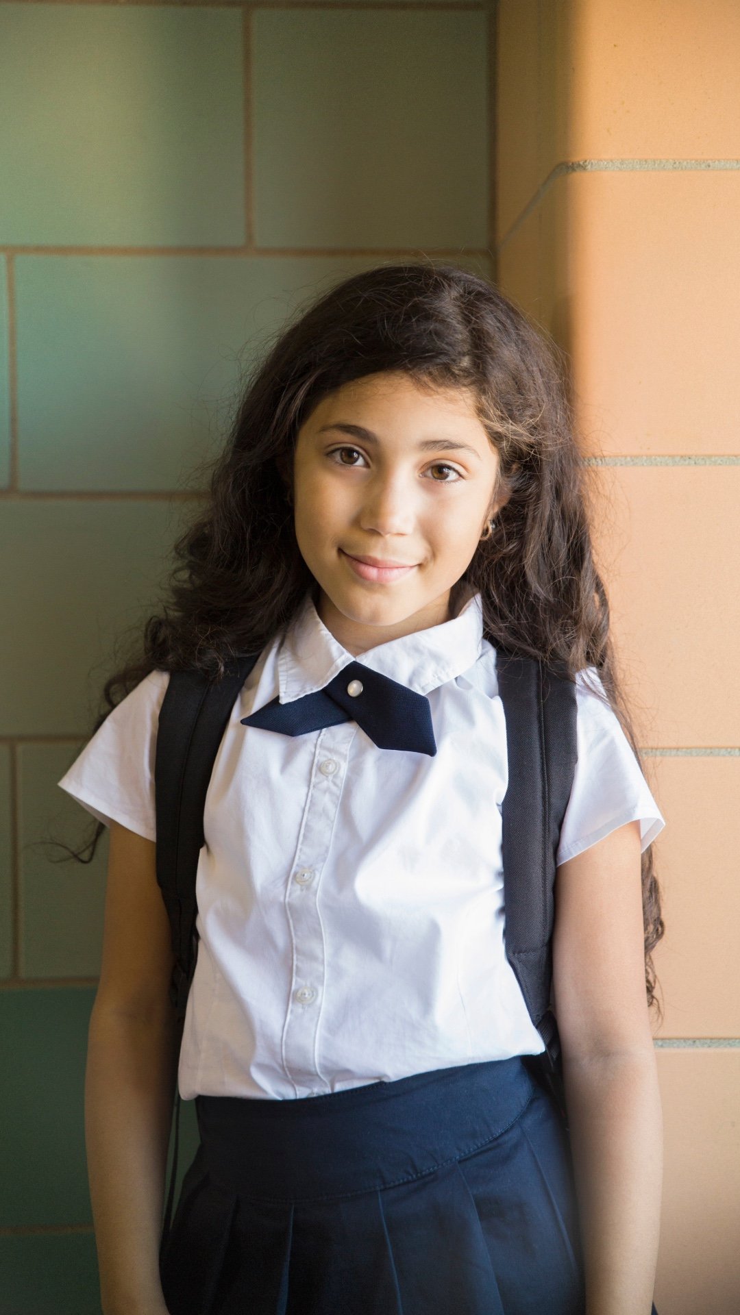 Young girl with long dark hair wearing a white shirt, dark blue skirt, and a navy blue bow tie, standing against a green and tan brick wall, carrying a black backpack.