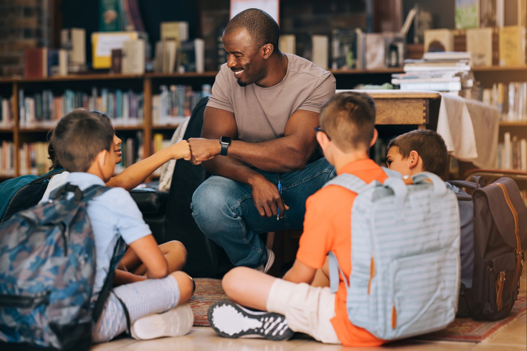 A man sitting on the floor in a library with four young boys, one of whom is shaking his fist to greet him.