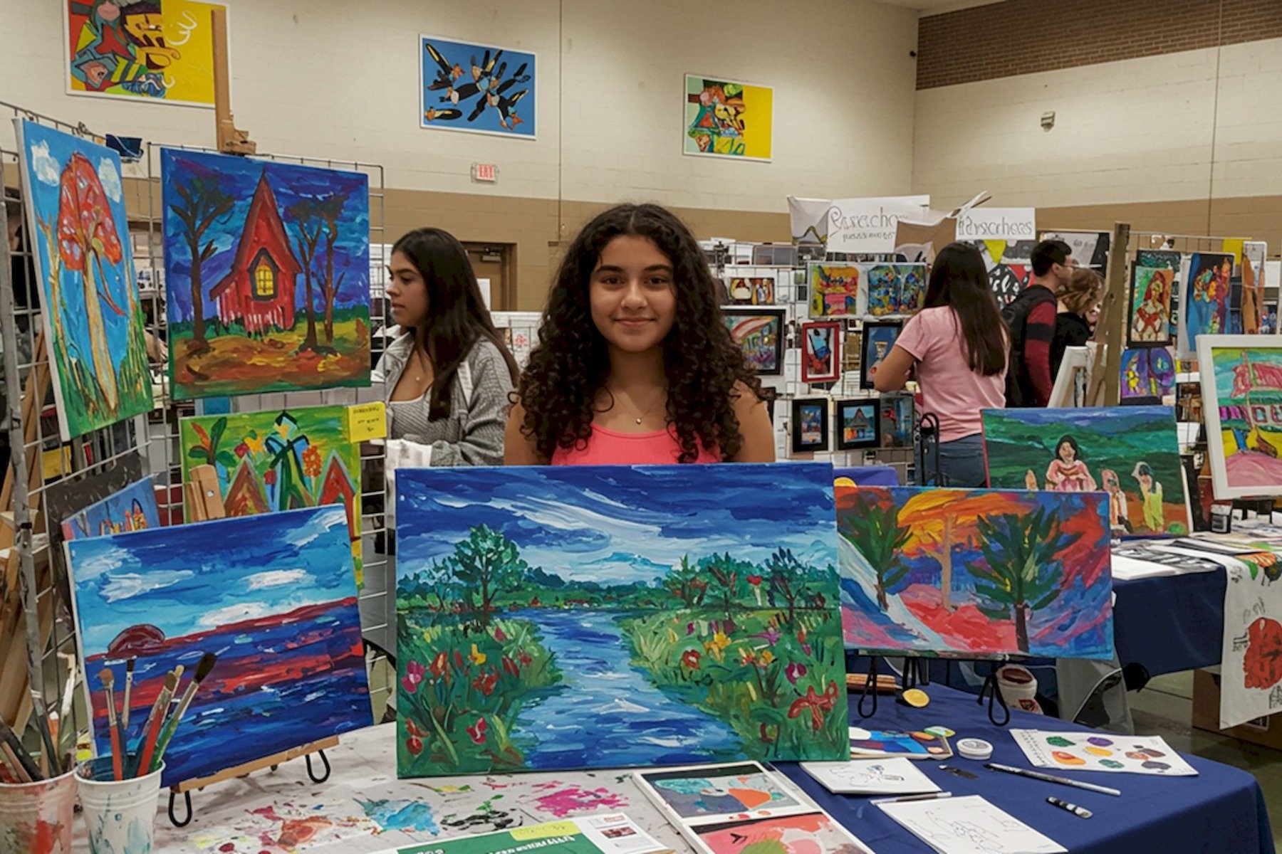 A girl with curly hair standing behind a table at an art fair displaying colorful landscape paintings, with other artists and their artwork visible in the background.