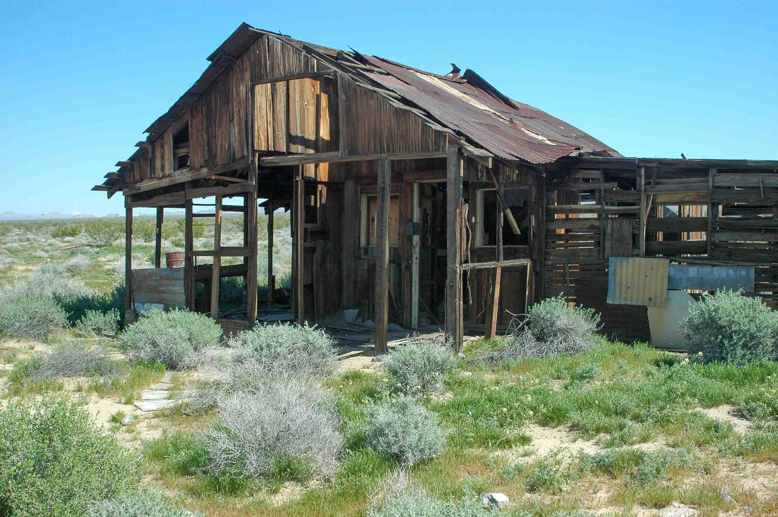 Old, dilapidated wooden house in a desert landscape with sparse bushes and clear blue sky.