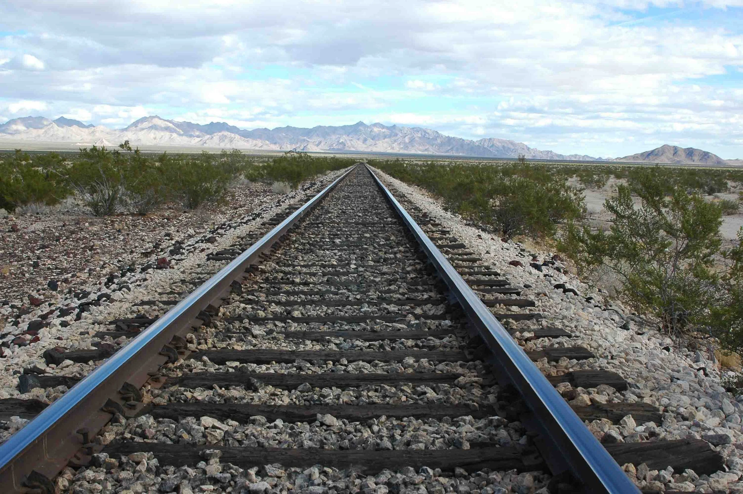 Train tracks running through a desert landscape with mountains in the distance, under a partly cloudy sky.