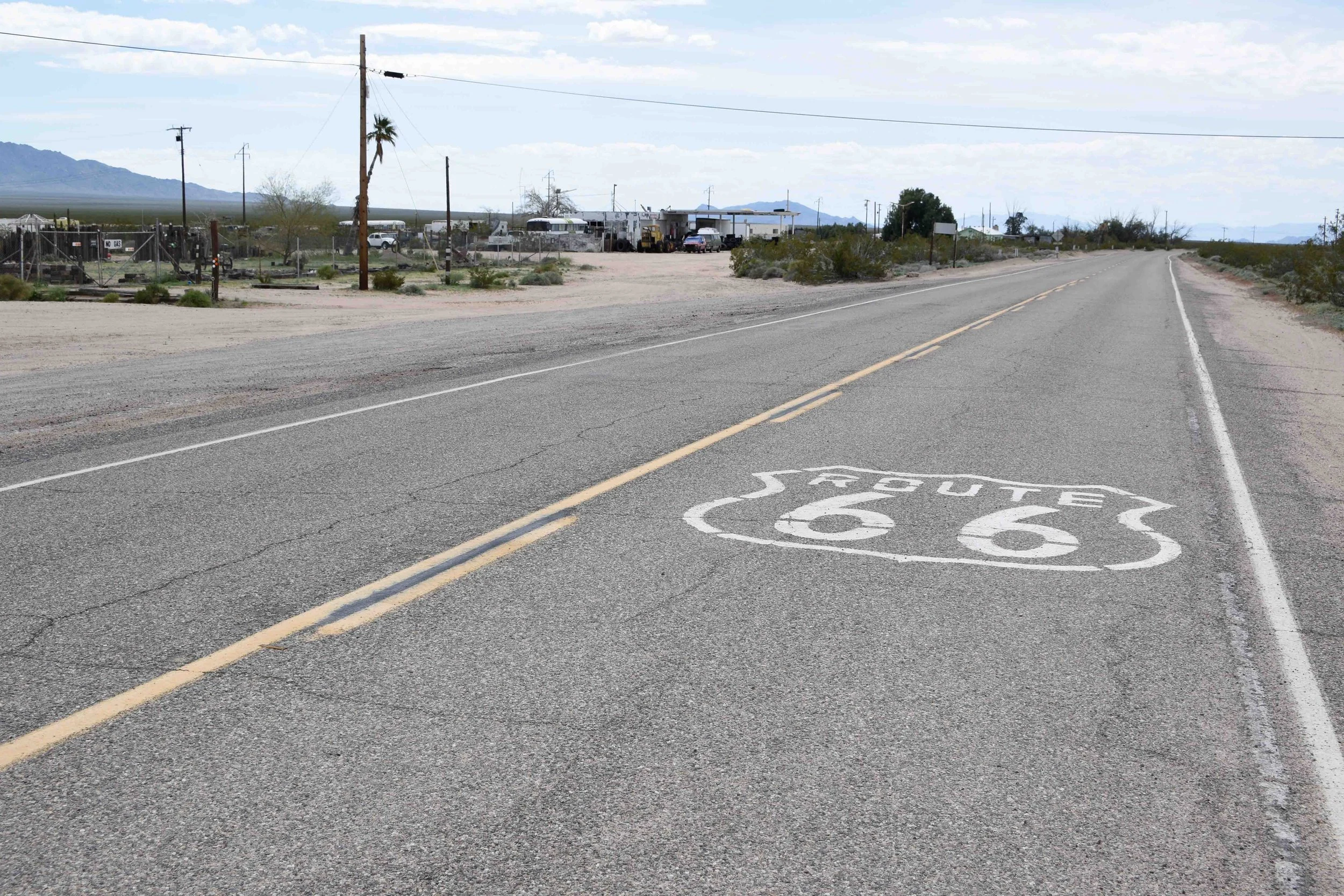 Desert highway with Route 66 shield painted on the road surface and a small roadside building in the background, under a cloudy sky.