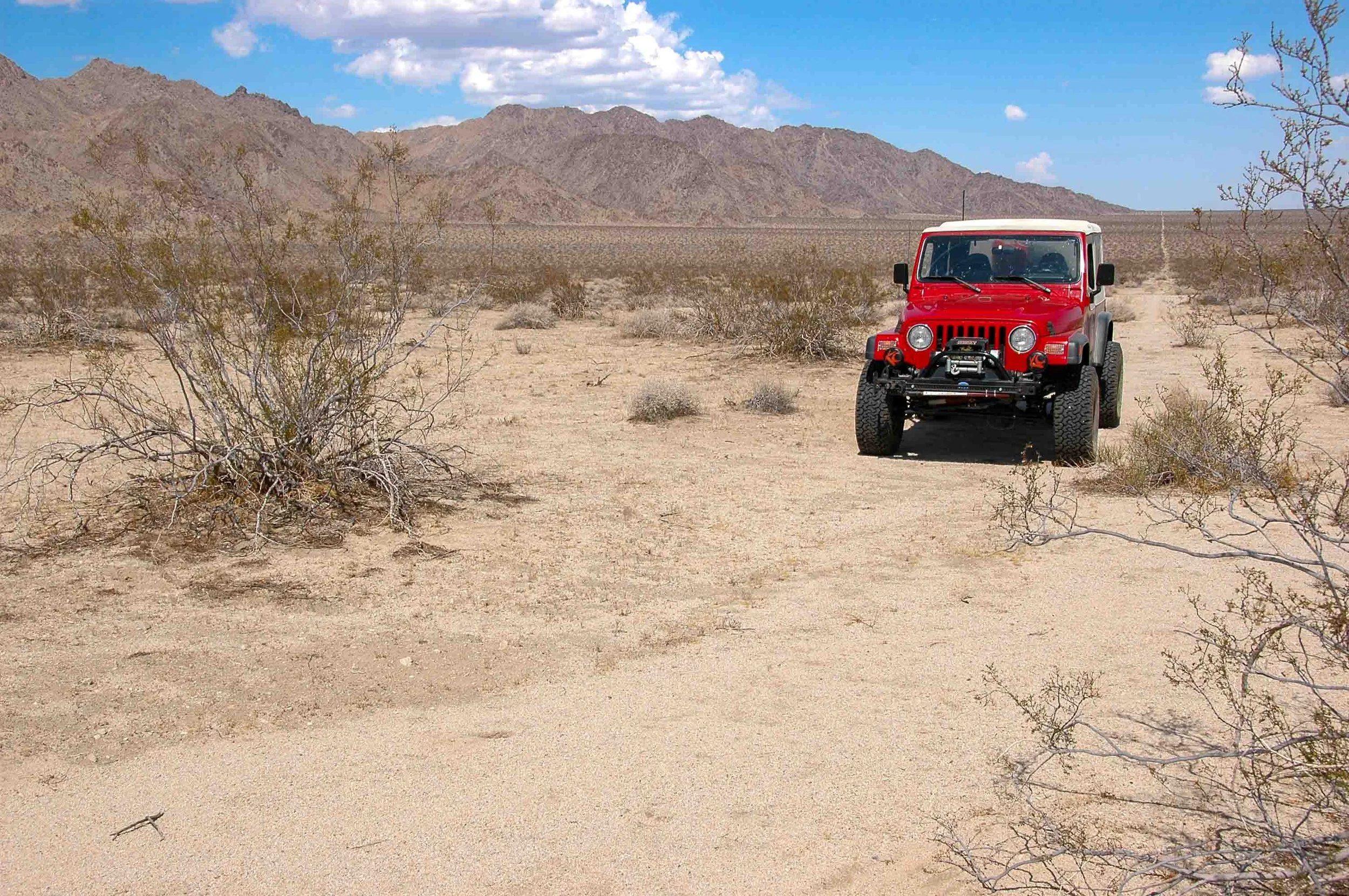 A red off-road vehicle parked in a desert landscape with sparse bushes, mountains in the background, and a partly cloudy sky.