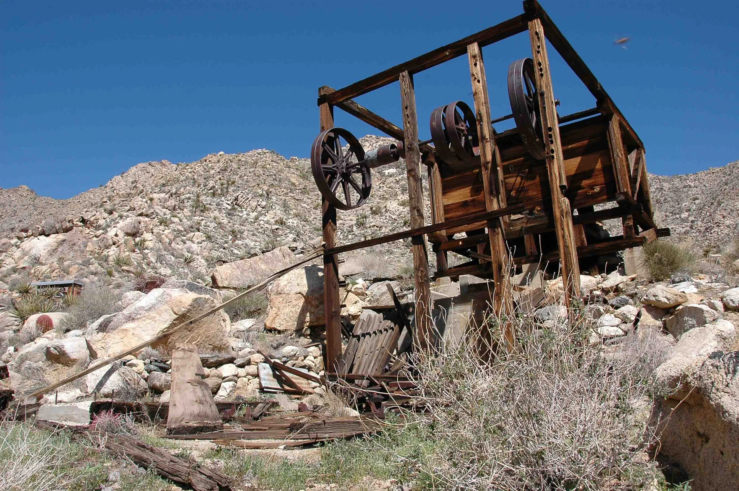 Old, abandoned wooden watermill with large metal wheels in a desert landscape with rocky terrain and sparse shrubs under a clear blue sky.