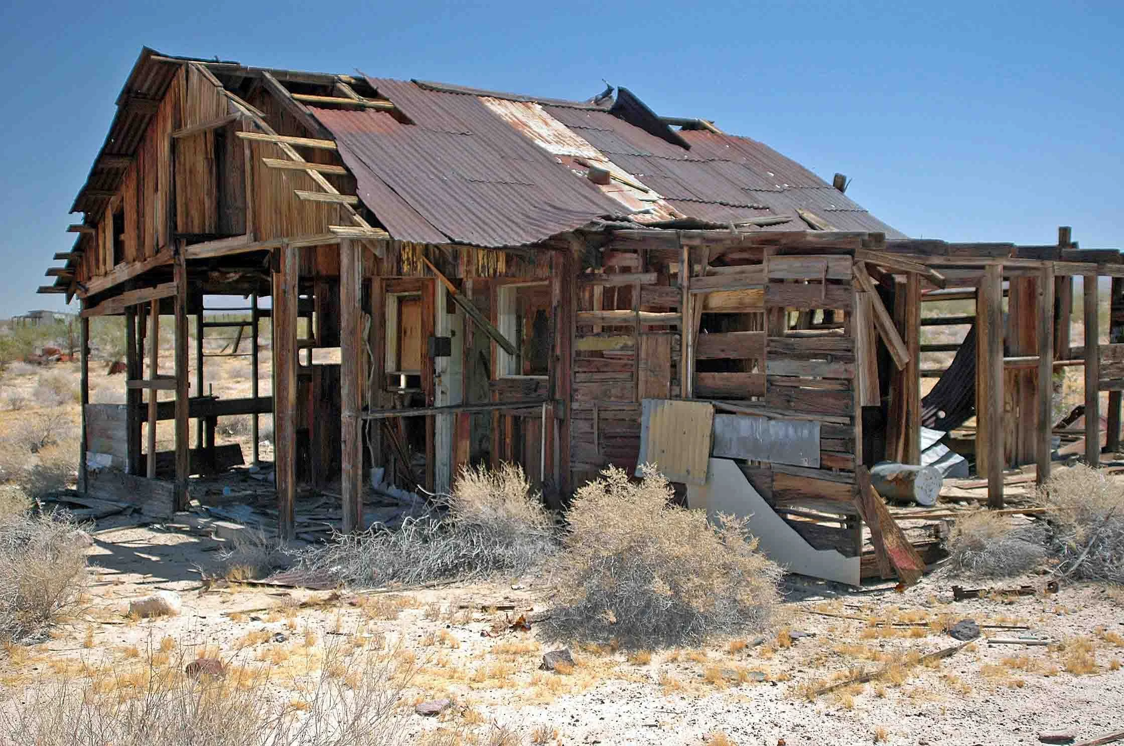 Dilapidated wooden house with a rusted metal roof in a desert landscape.