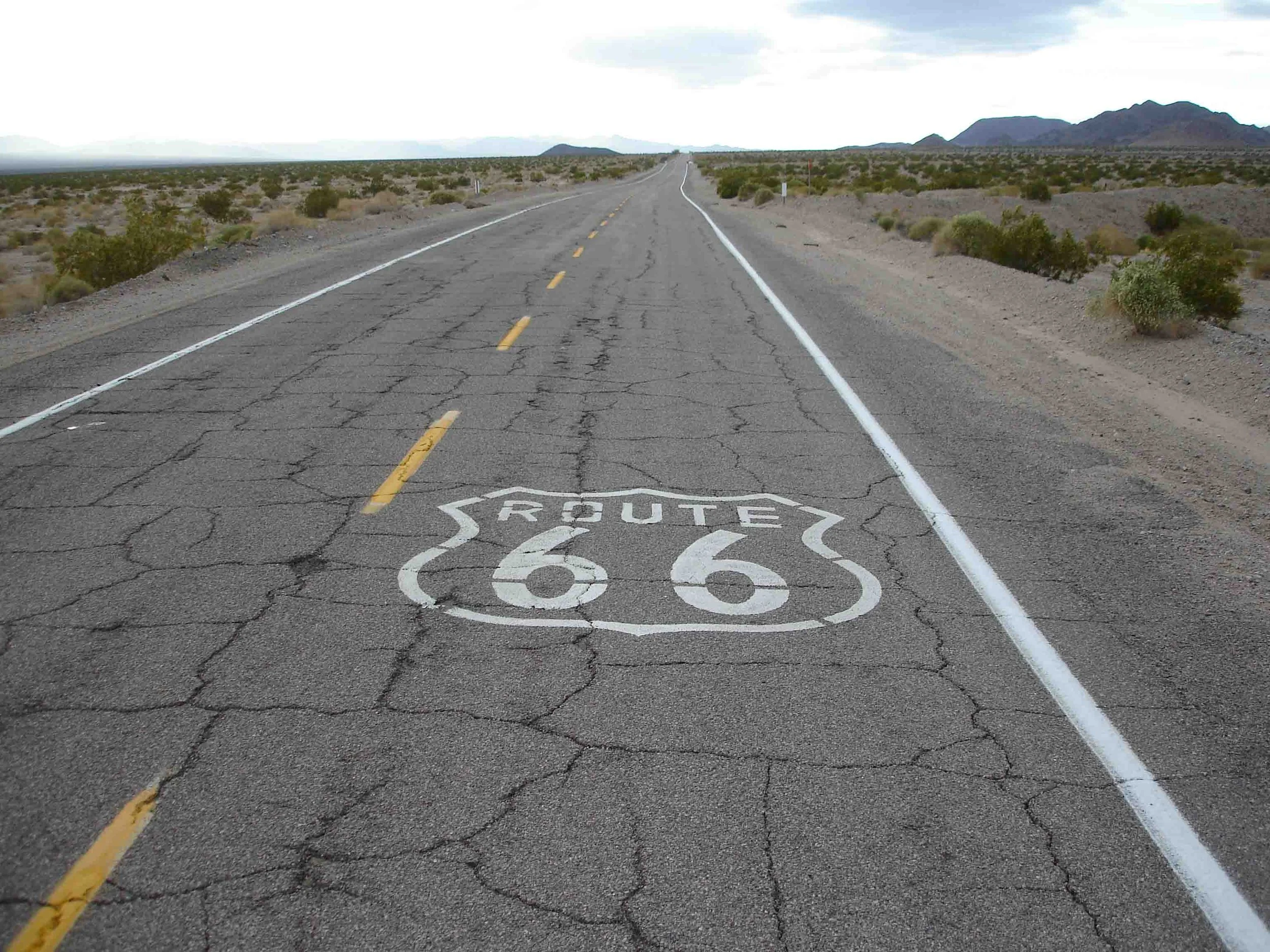 Long stretch of Route 66 highway in a desert landscape with cracked asphalt and sparse vegetation, with distant mountains under a cloudy sky.