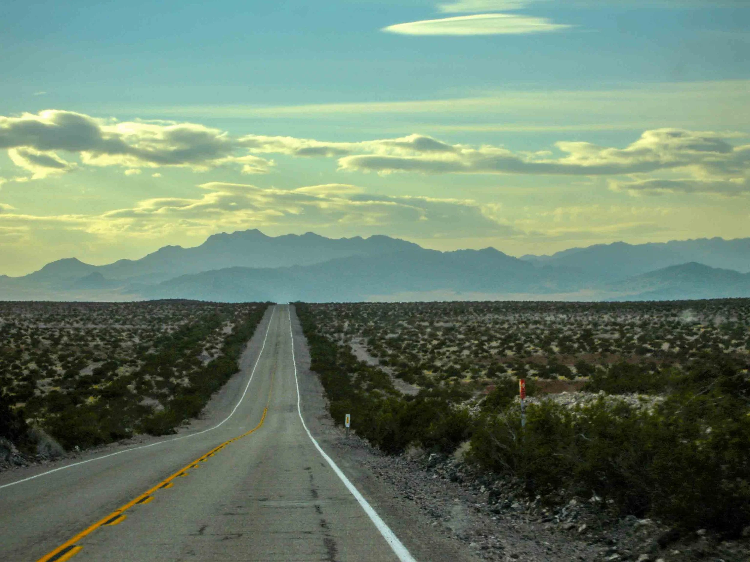 A straight road extending into the distance through a desert landscape with sparse vegetation, with mountains and a partly cloudy sky in the background.
