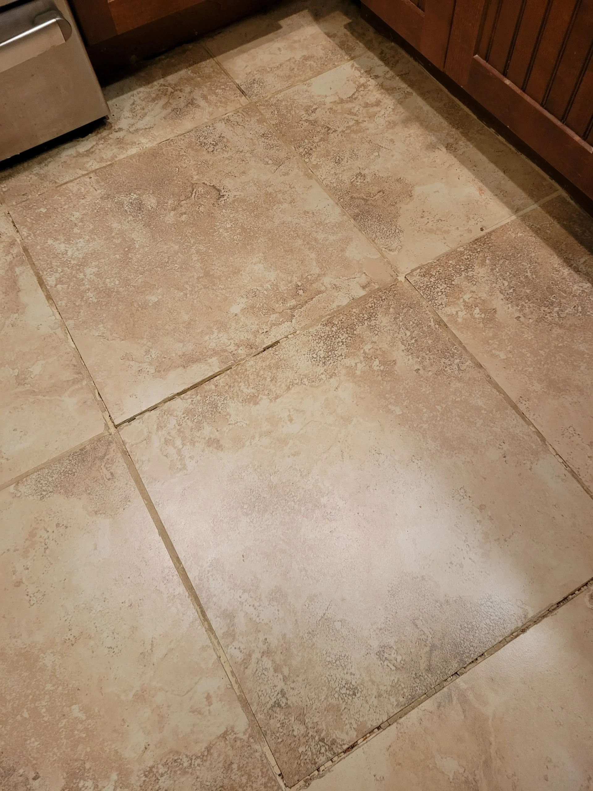 A close-up of a beige tiled kitchen floor with large square tiles showing textured patterns and grout lines.