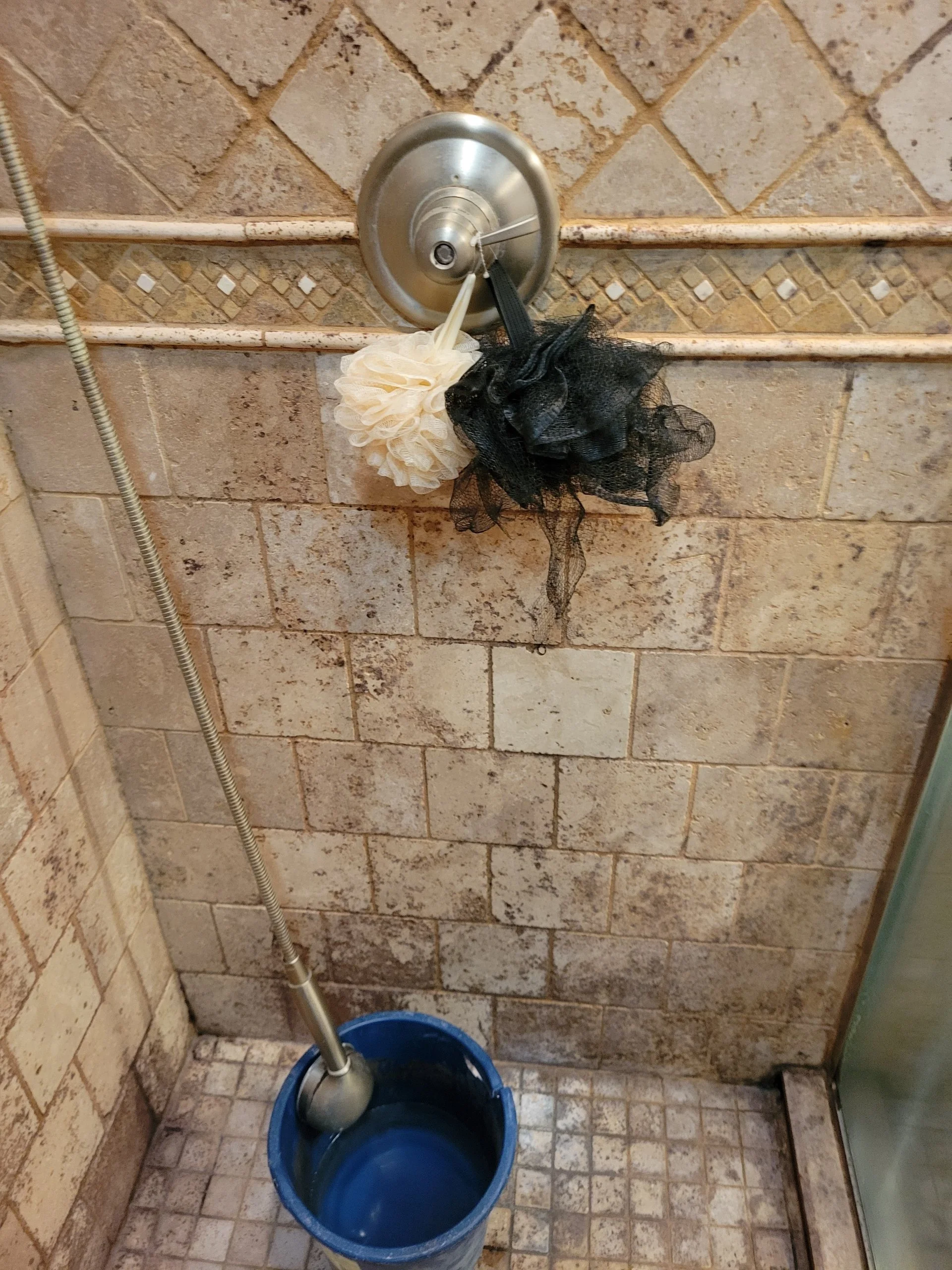 Bathroom shower with beige tile walls, a metal shower head, and a blue bucket on the floor containing a shower tool. Two bath loofahs, one white and one black, hang from the shower head.