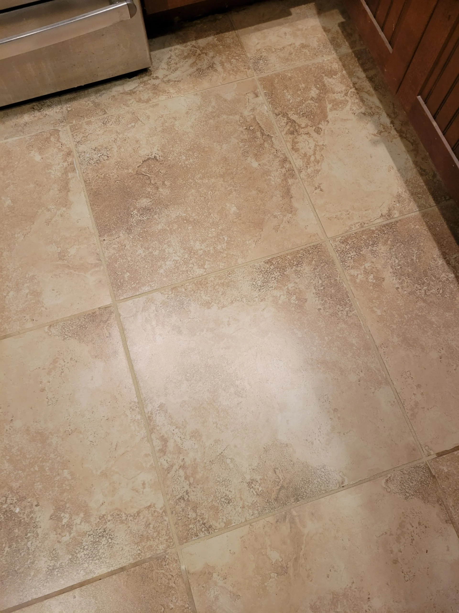 Tile floor with a beige and brown textured pattern, next to a stainless steel appliance and wooden cabinet.