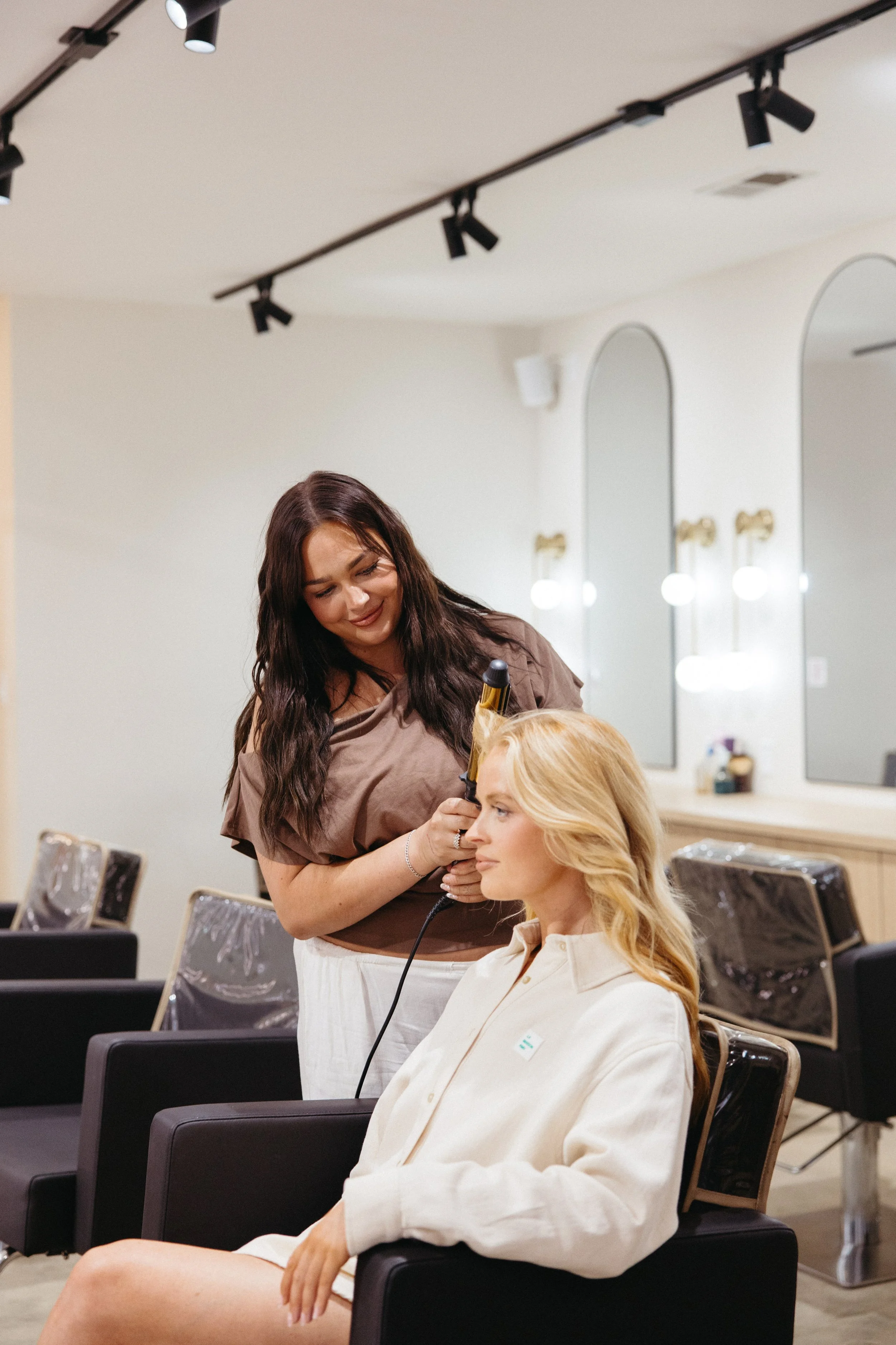A hairstylist with long dark hair styling a blonde woman's hair in a salon mirror. The blonde woman is wearing a cream-colored robe and sitting in a salon chair. The salon is bright with modern decor and mirror lights.