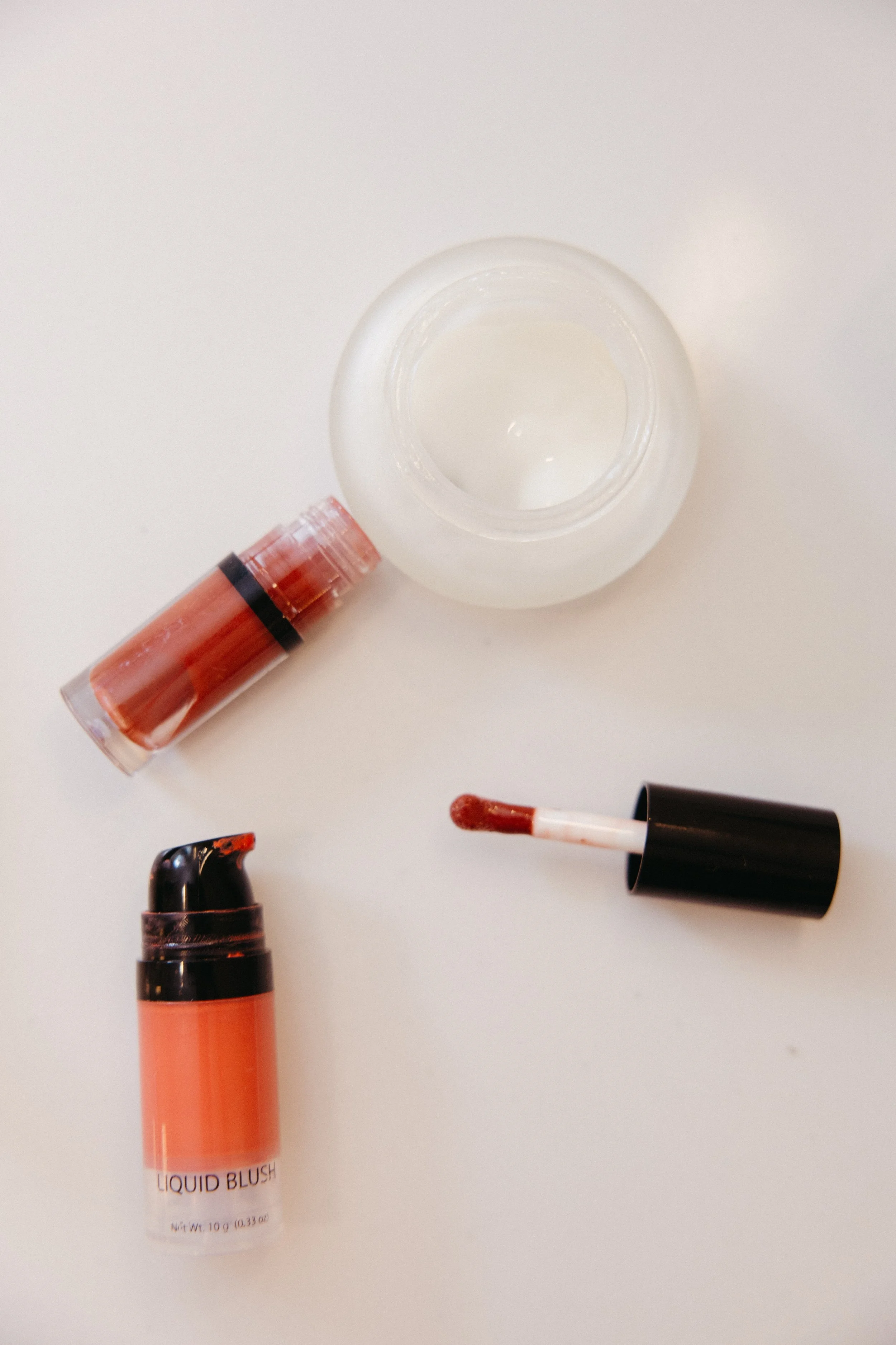 Various makeup products on a white surface, including a liquid blush, a lip gloss, and a lipstick with an applicator stick.