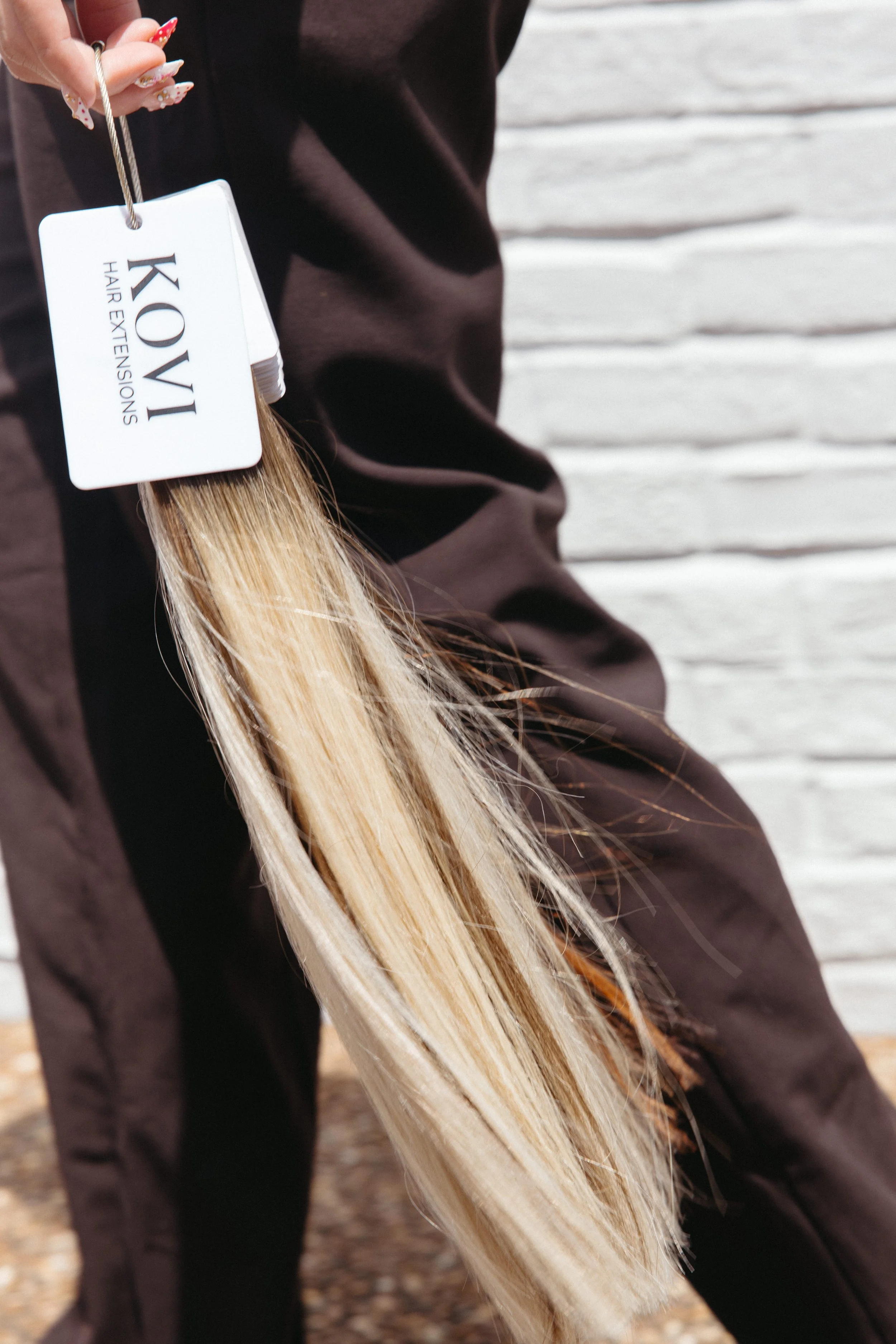 A person holding a hair extension sample with long, straight, blonde hair and a "KOVI Hair Extensions" tag, standing outdoors.