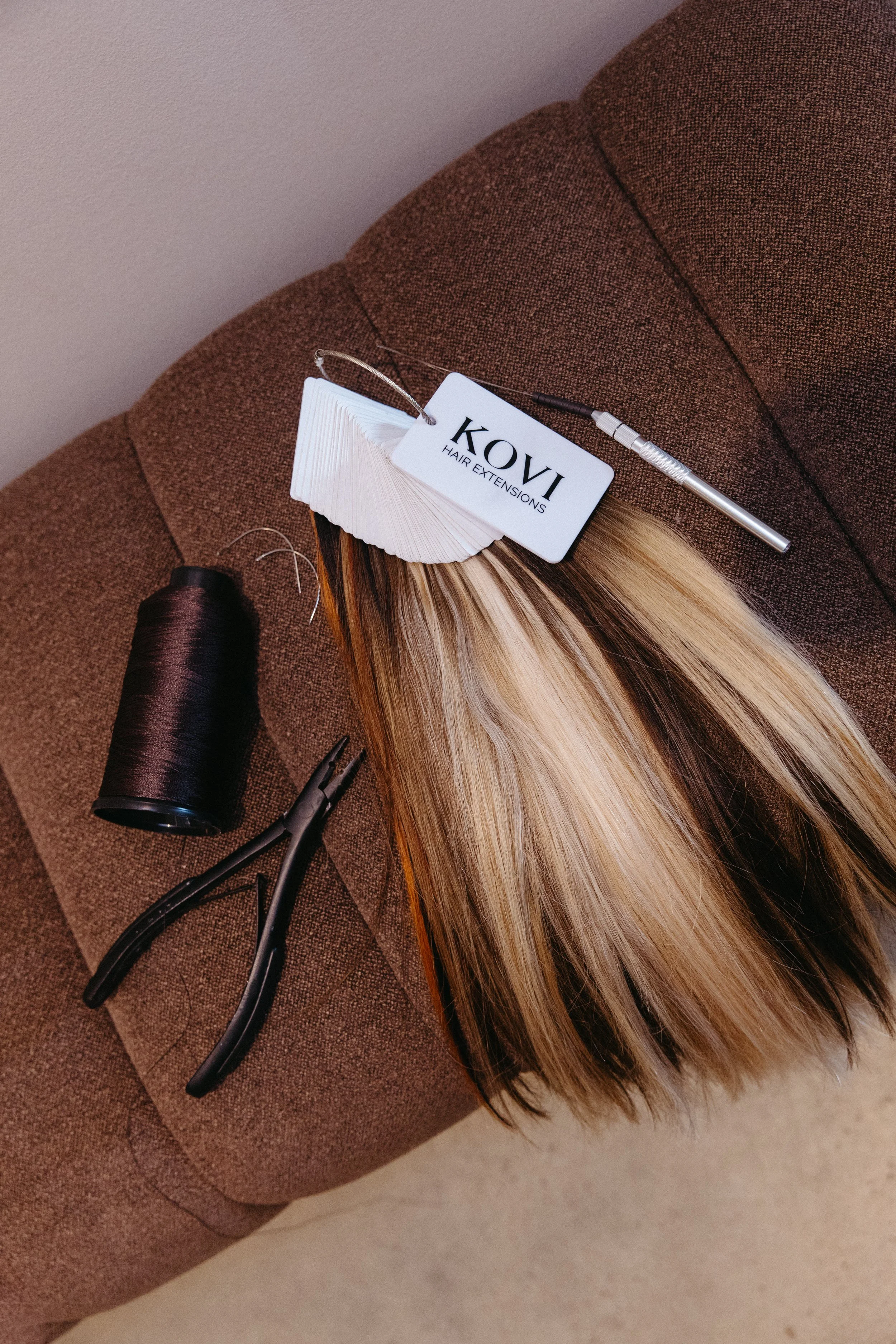 Sample swatch book for KOVI Hair Extensions on a brown textured surface, with a bundle of blonde hair extensions, a spool of black thread, and a pair of black scissors nearby.