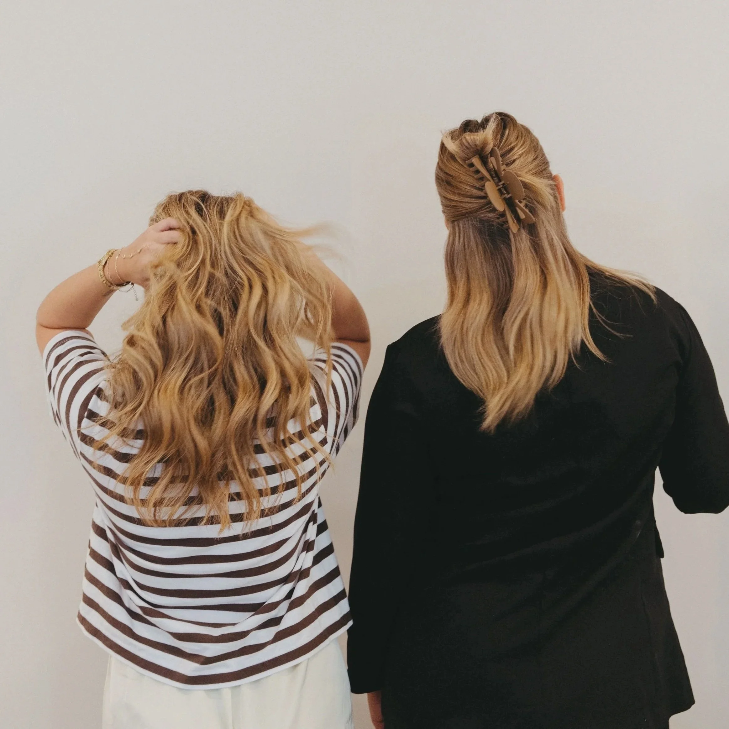 Two women standing with their backs to the camera, one with curly blonde hair wearing a striped shirt and white pants, the other with straight blonde hair secured with hair clips, wearing a black top and white skirt, against a plain light-colored wall.