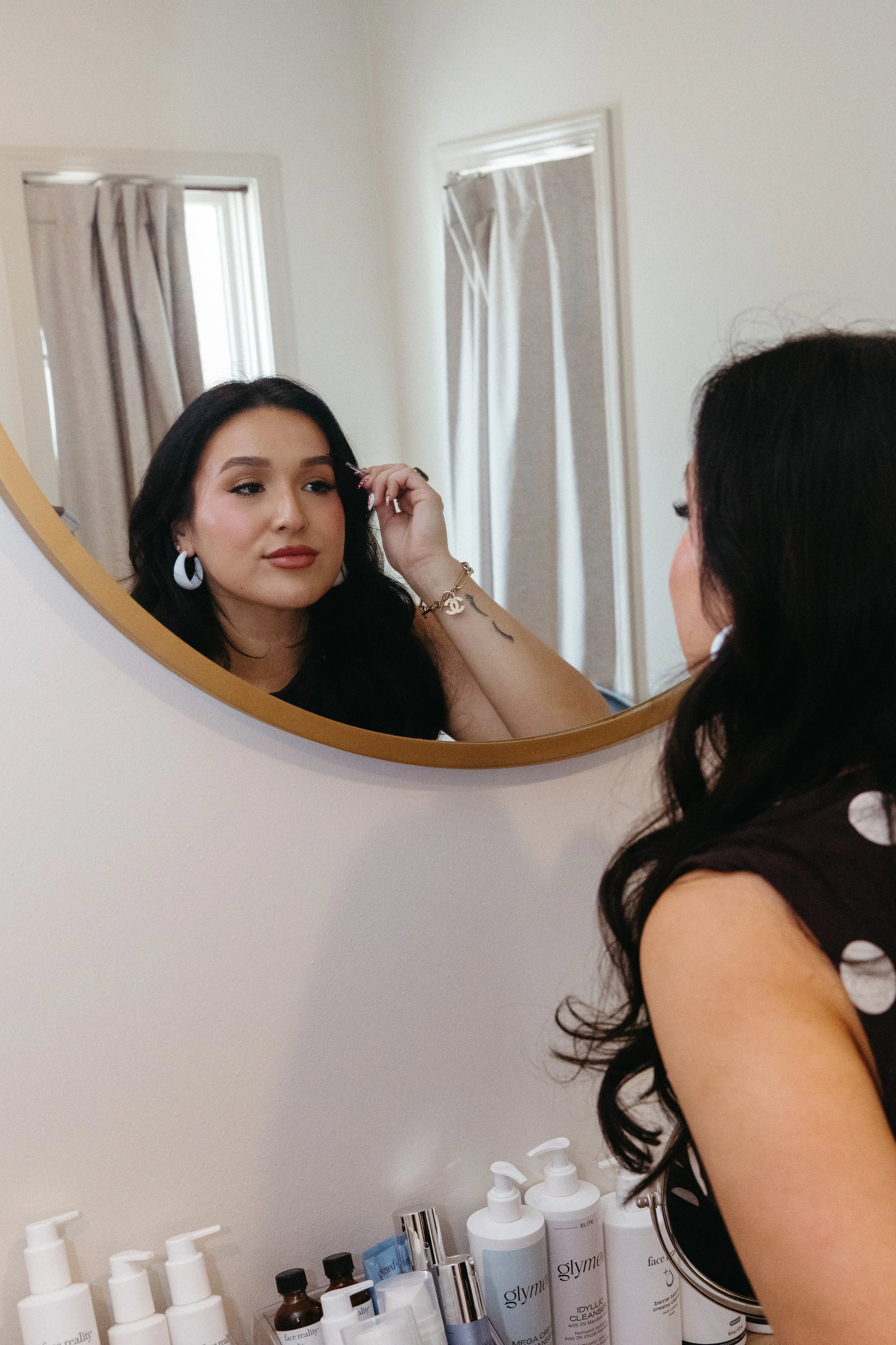 A woman with long dark hair applying makeup or adjusting her eyelashes in a bathroom mirror.