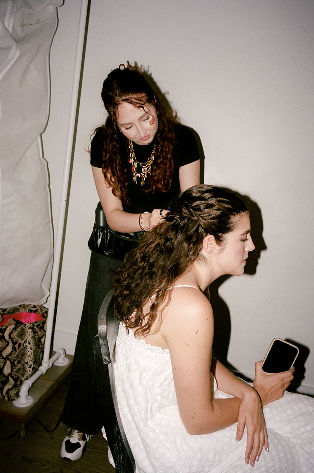 A woman with curly brown hair is sitting in a white dress, looking at her phone. A stylist with long, curly hair is standing behind her, braiding her hair.