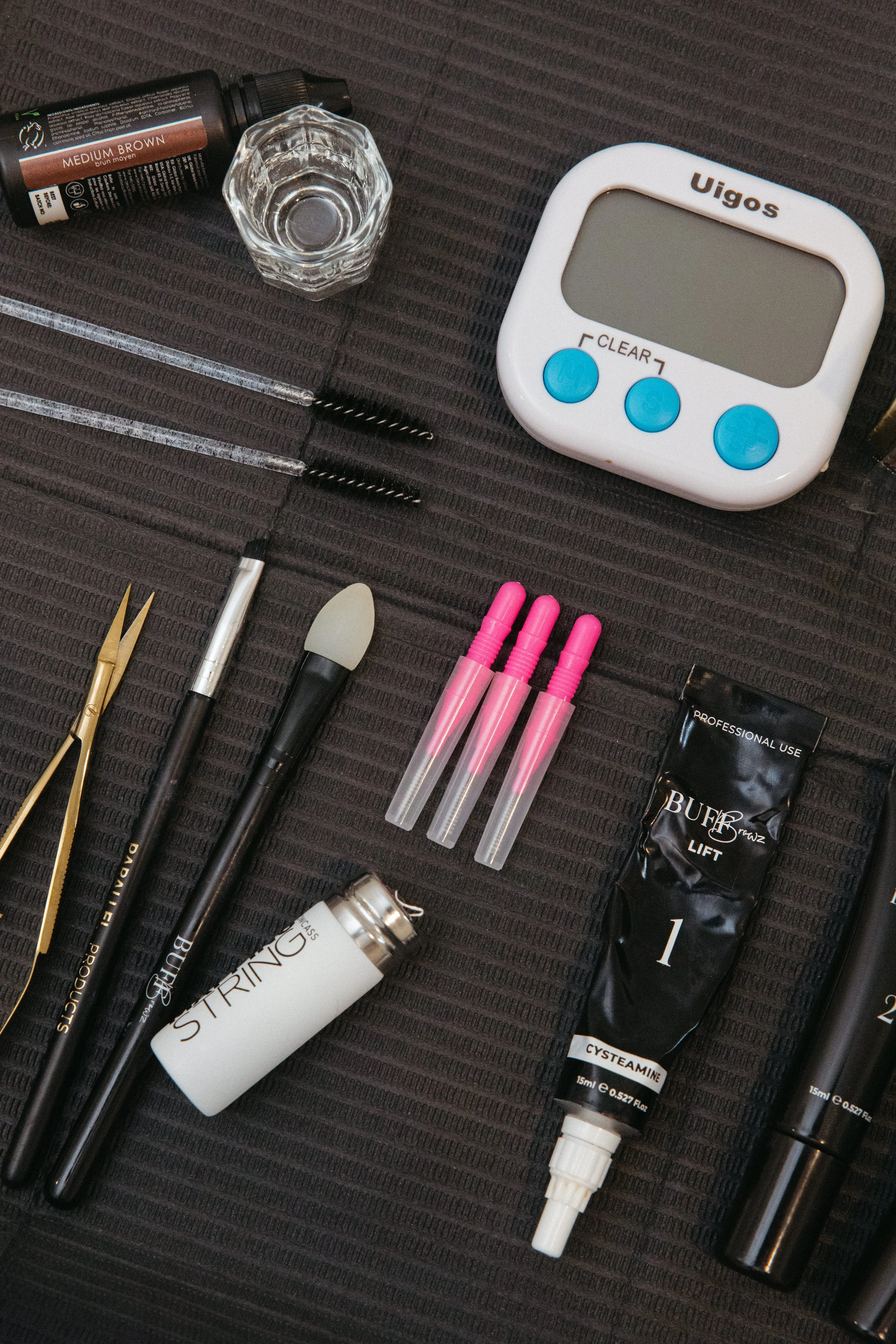 A collection of beauty and skincare products, including tubes of various creams, serums, a white electronic device labeled 'Uigos', small skincare bottles with pink caps, a pair of tweezers, a black tube, a small glass cup, and makeup brushes on a black textured surface.