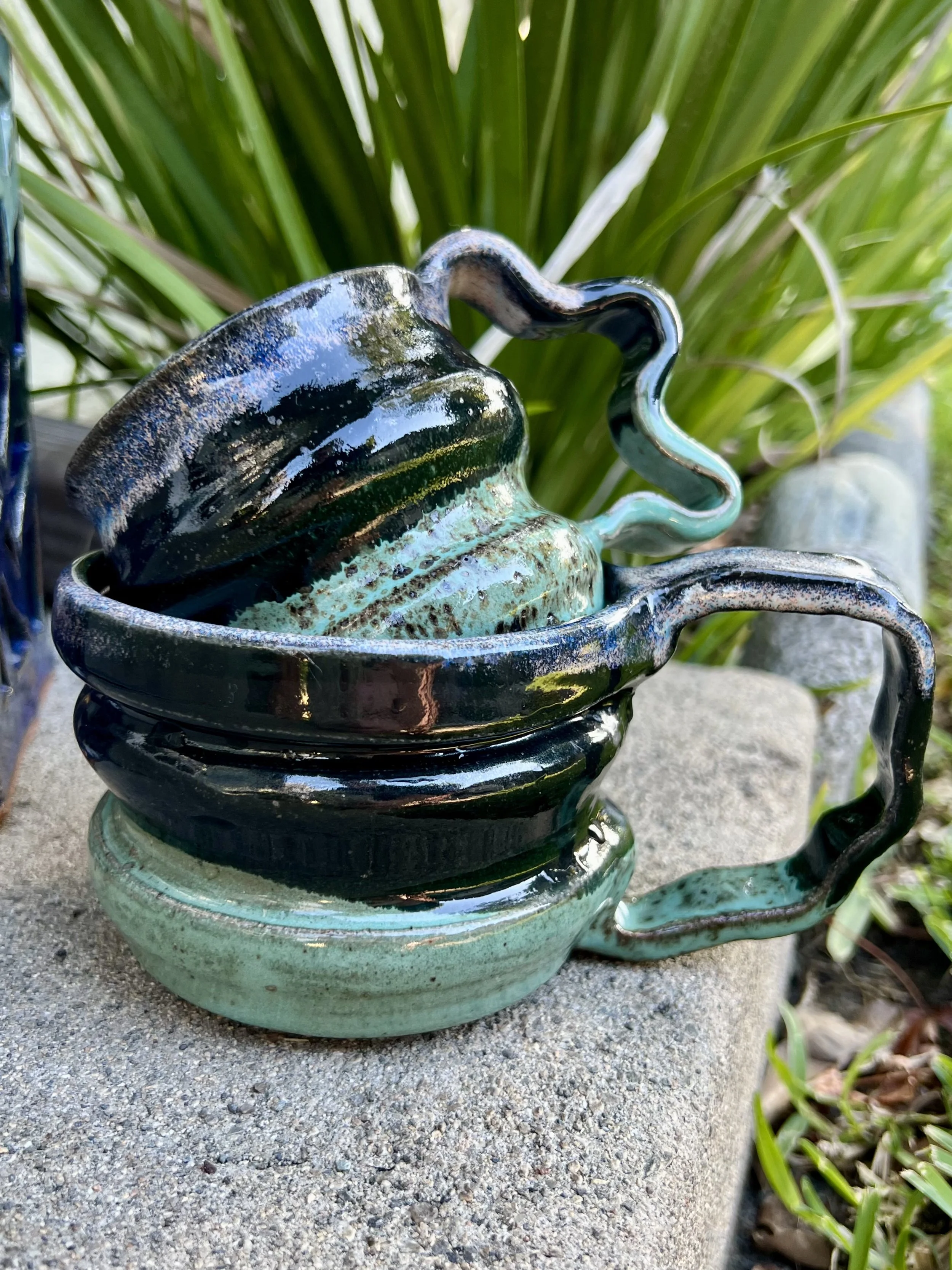 Two decorative ceramic mugs with wavy handles, glazed with earthy blacks and greens