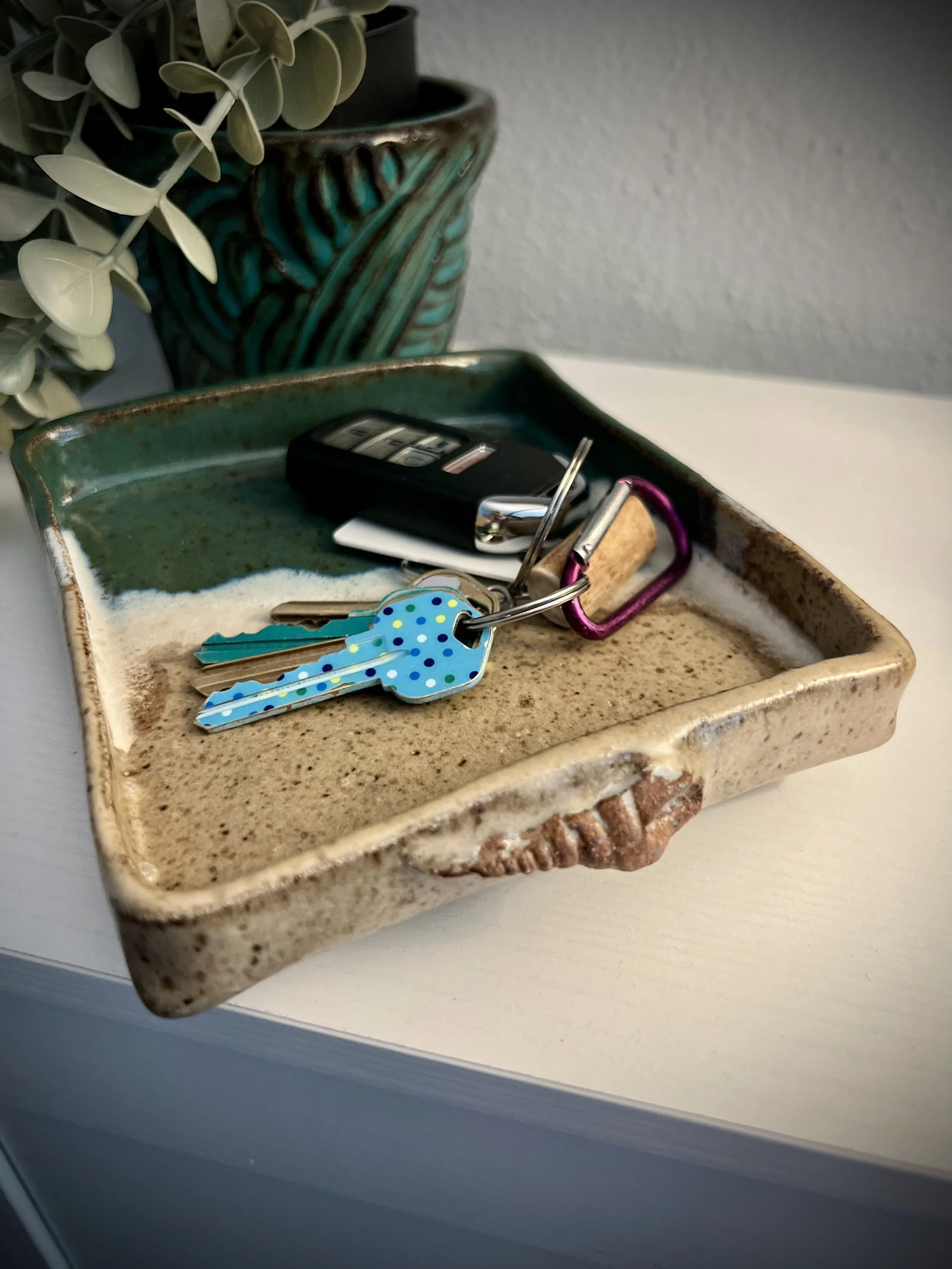 A ceramic tray with a sand-colored base contains a keyring with keys, a car key fob, and a pen.