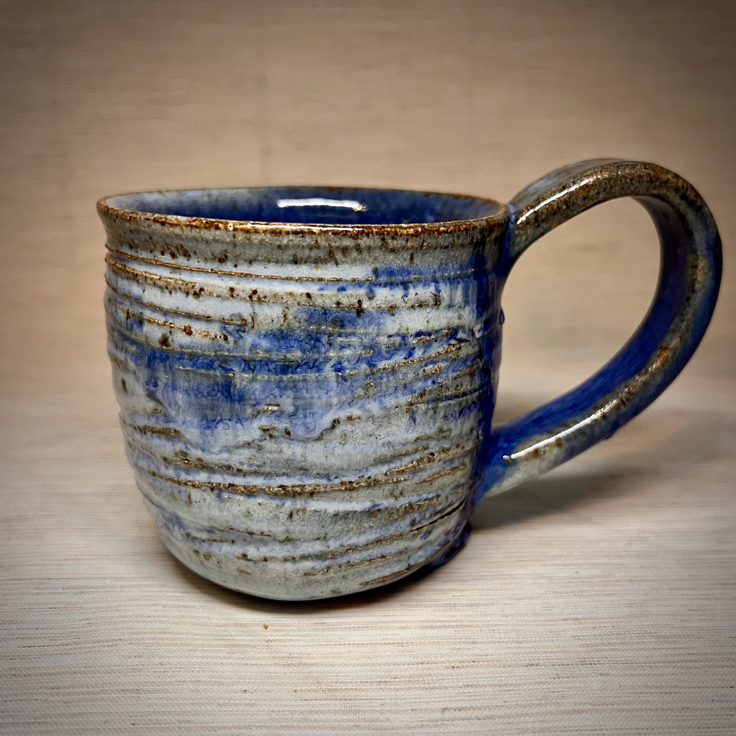 A handmade ceramic mug with a textured, ribbed surface, glazed in shades of blue mixed with natural clay.
