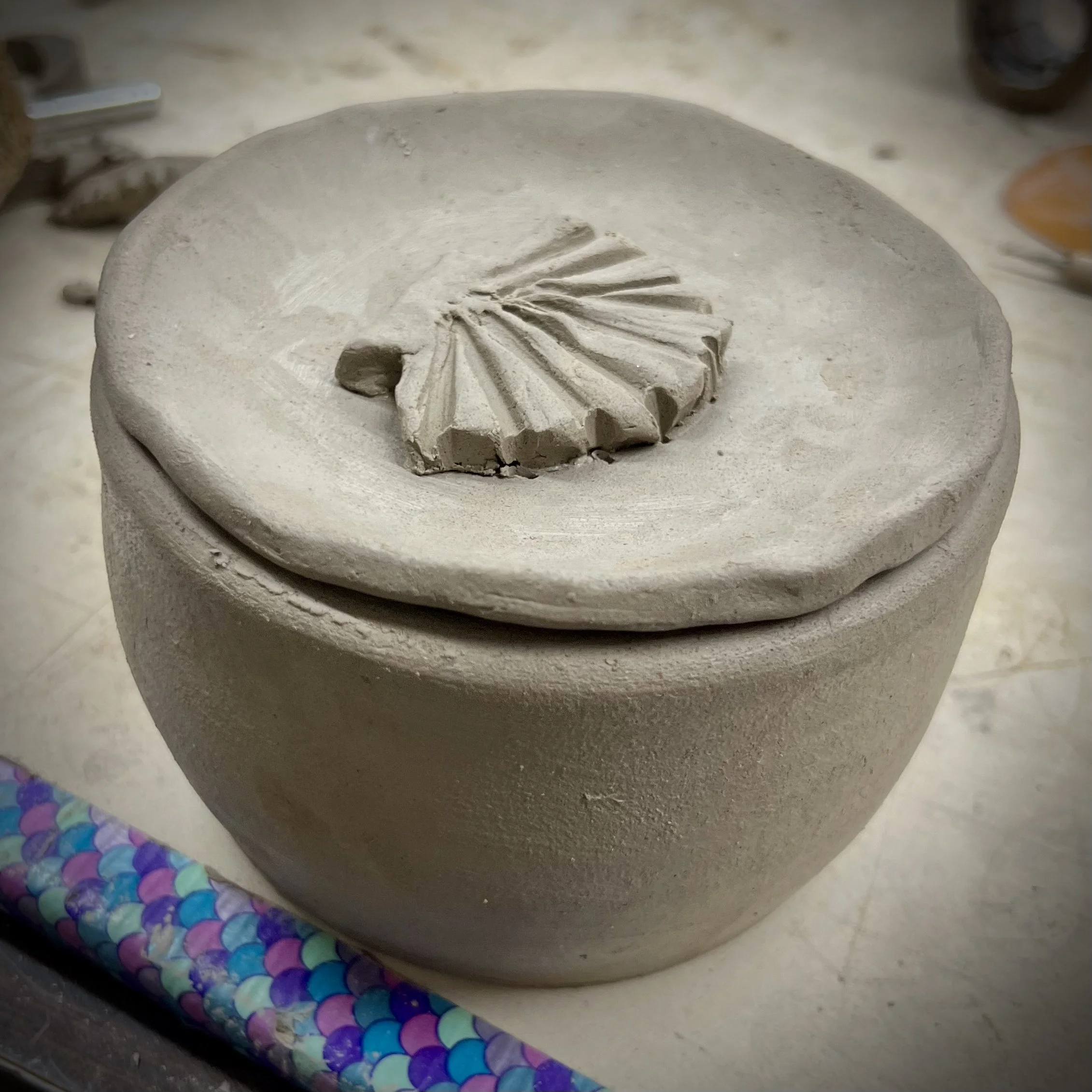 A round, unfinished clay pot with a spiral carved design on the lid, placed on a table with a colorful, patterned rolling pin nearby.