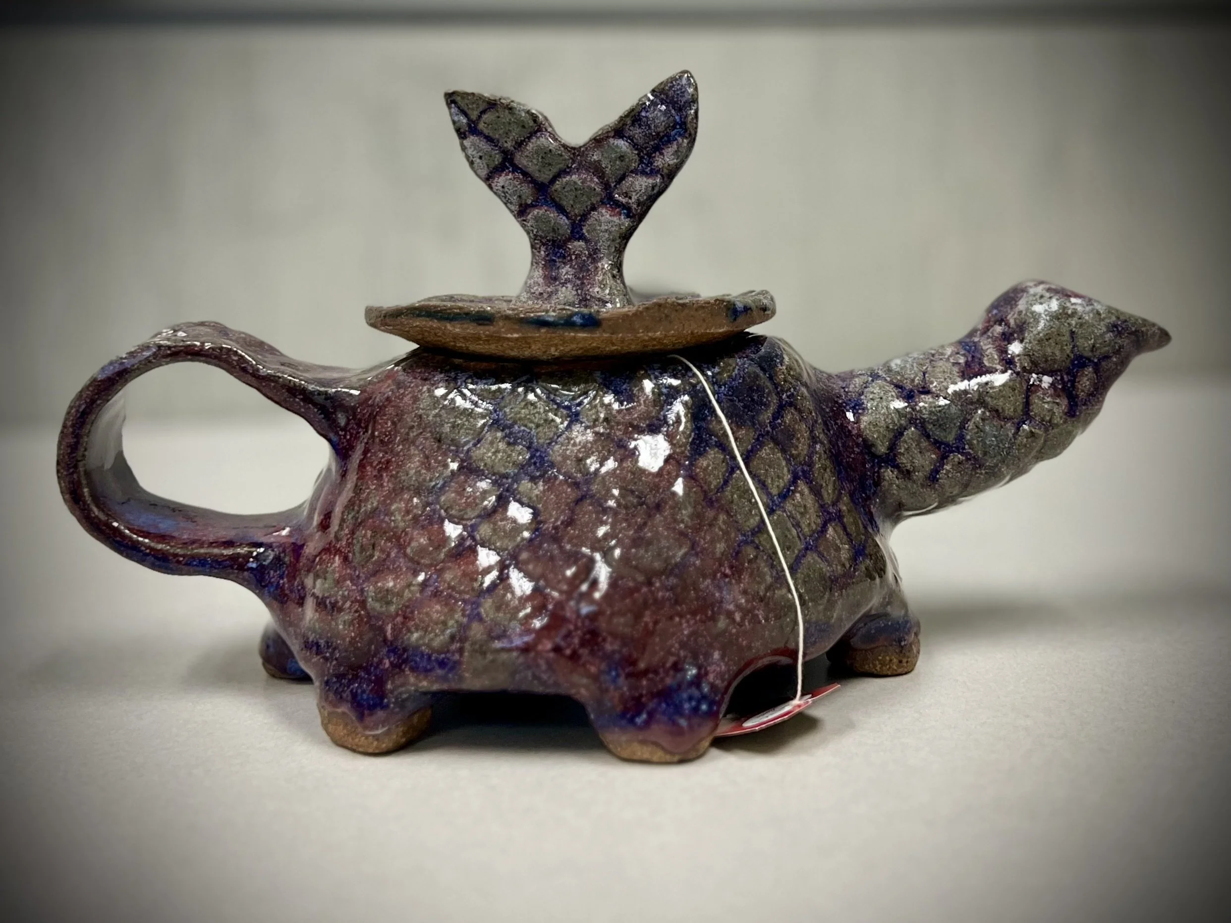 A ceramic teapot with a unique design, a textured surface, iridescent glaze, a handle on the side, a spout, and a small whale-shaped figure on the lid.