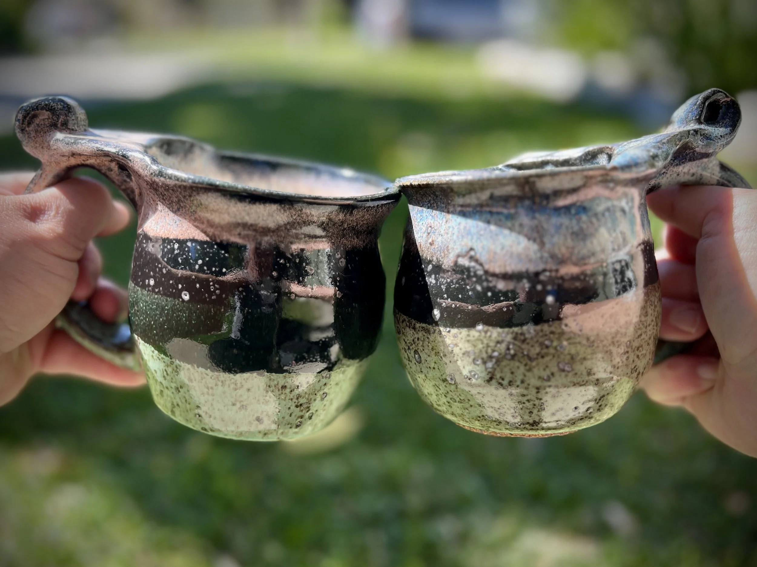 Two ceramic mugs, hand-crafted with a earthy black and green glaze finish, clinking together outdoors.