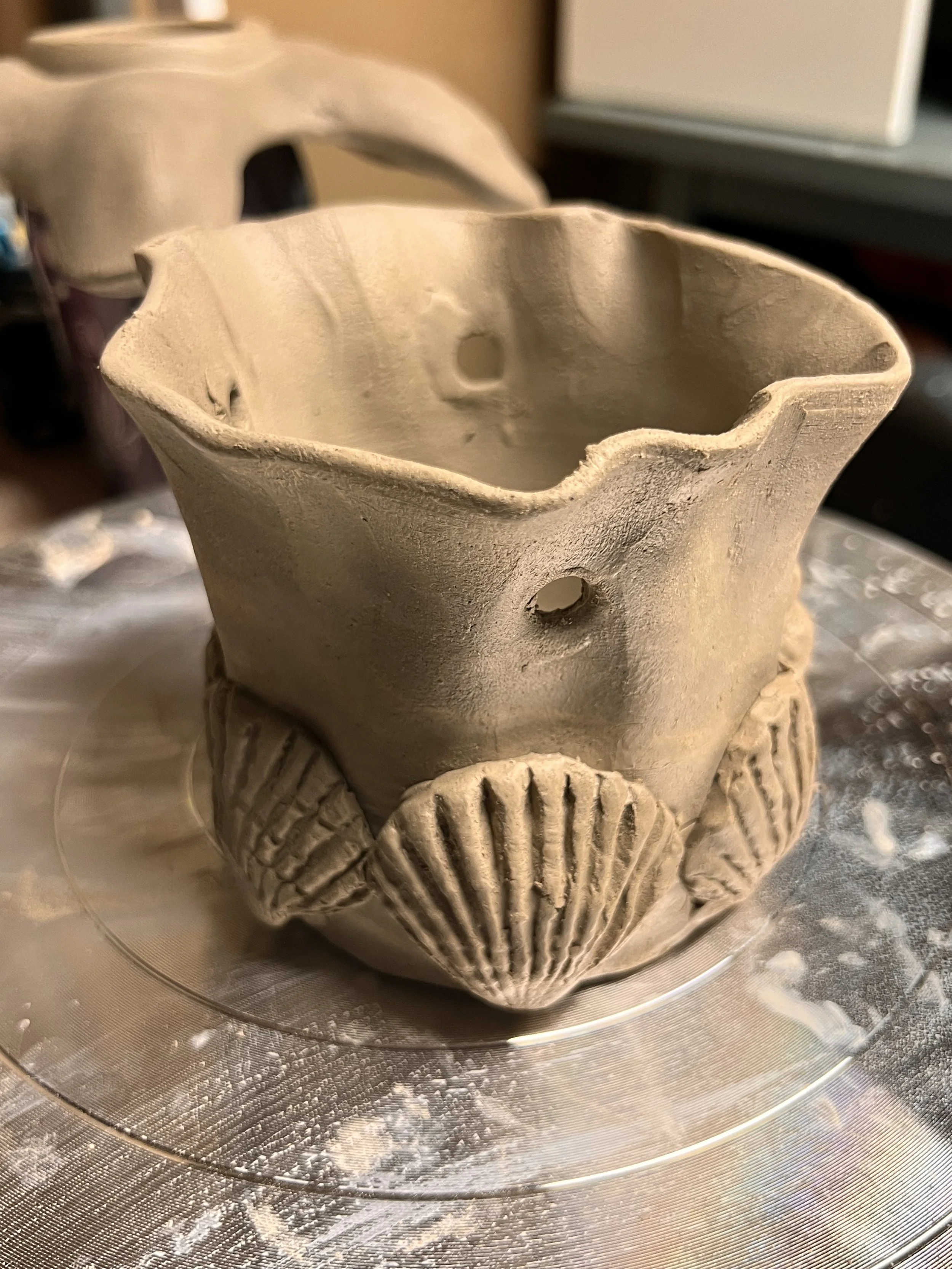 A ceramic flower pot with an irregular wavy rim, decorated with shell-shaped clay appliqués around the base, placed on a turntable or pottery wheel.