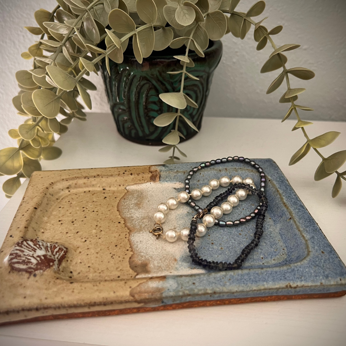 Decorative ceramic tray with three necklaces, a potted plant with trailing leaves in a dark green pot in the background.