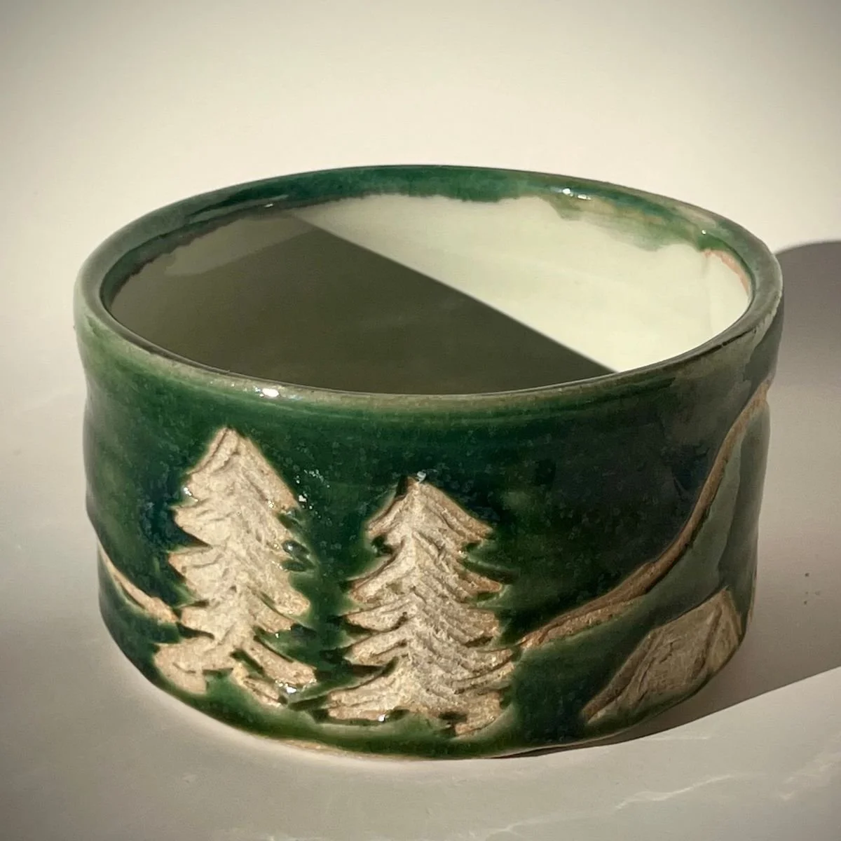 Ceramic bowl with a painted forest scene, featuring pine trees, placed on a flat surface with a shadow cast to its right.