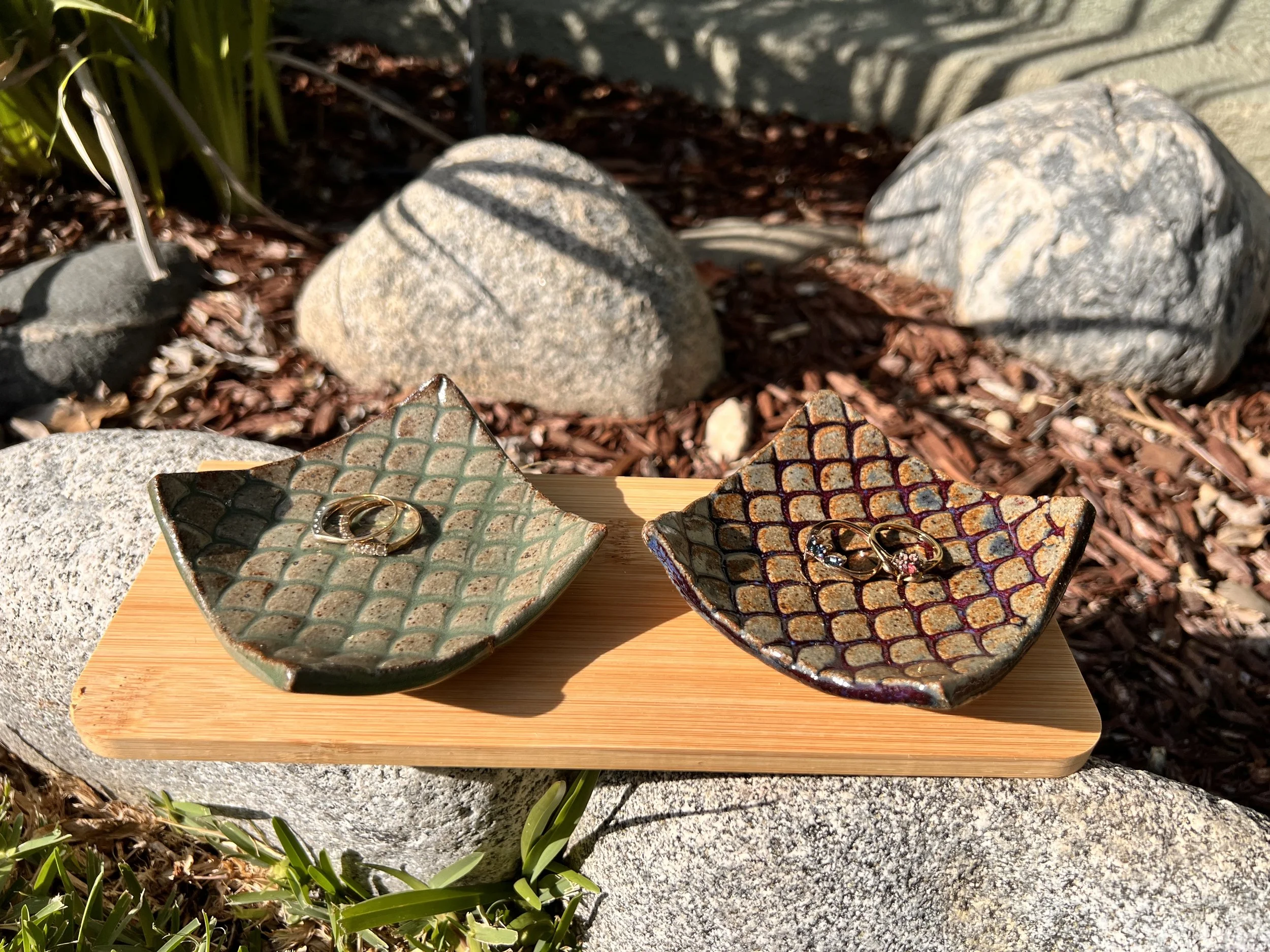Two ceramic dishes with geometric patterns and rings on top, placed on a wooden platform outdoors, surrounded by rocks and plants.