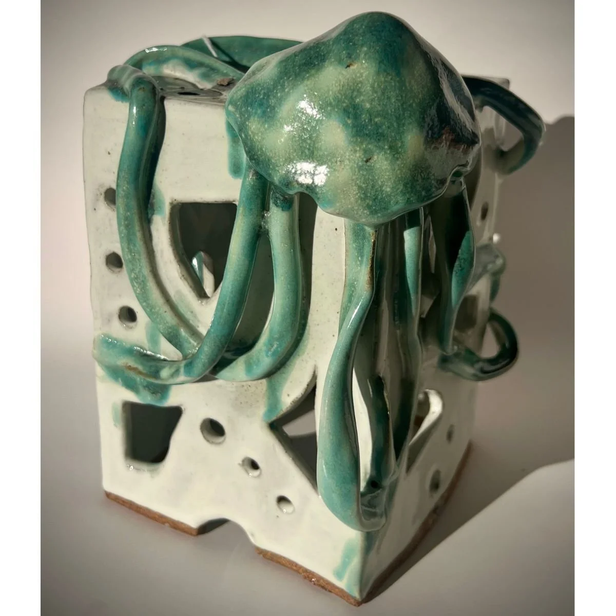 A ceramic sculpture of an octopus, featuring a cuboid base with multiple holes, with tentacles and head extending from the top, glazed in green, turquoise, and white colors.