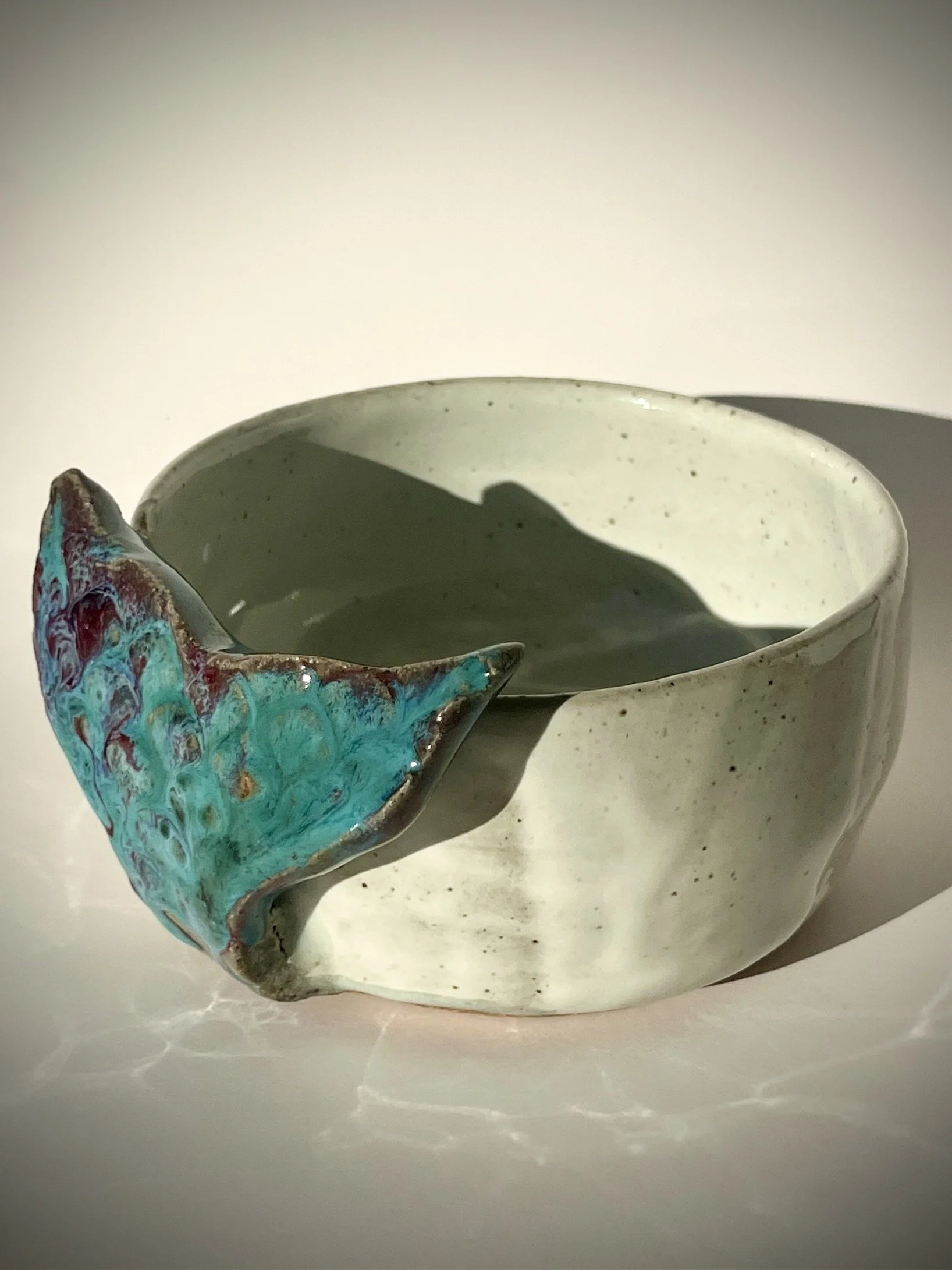 A white ceramic bowl with a textured surface, featuring a large turquoise and red glaze whale tail-shaped ornament on its side.