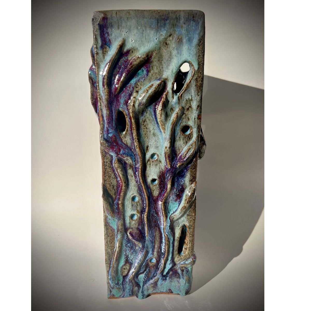 A ceramic sculpture with an abstract, organic design featuring flowing, curving lines and hollow holes, glazed in shades of blue, purple, and glaze with a textured finish.