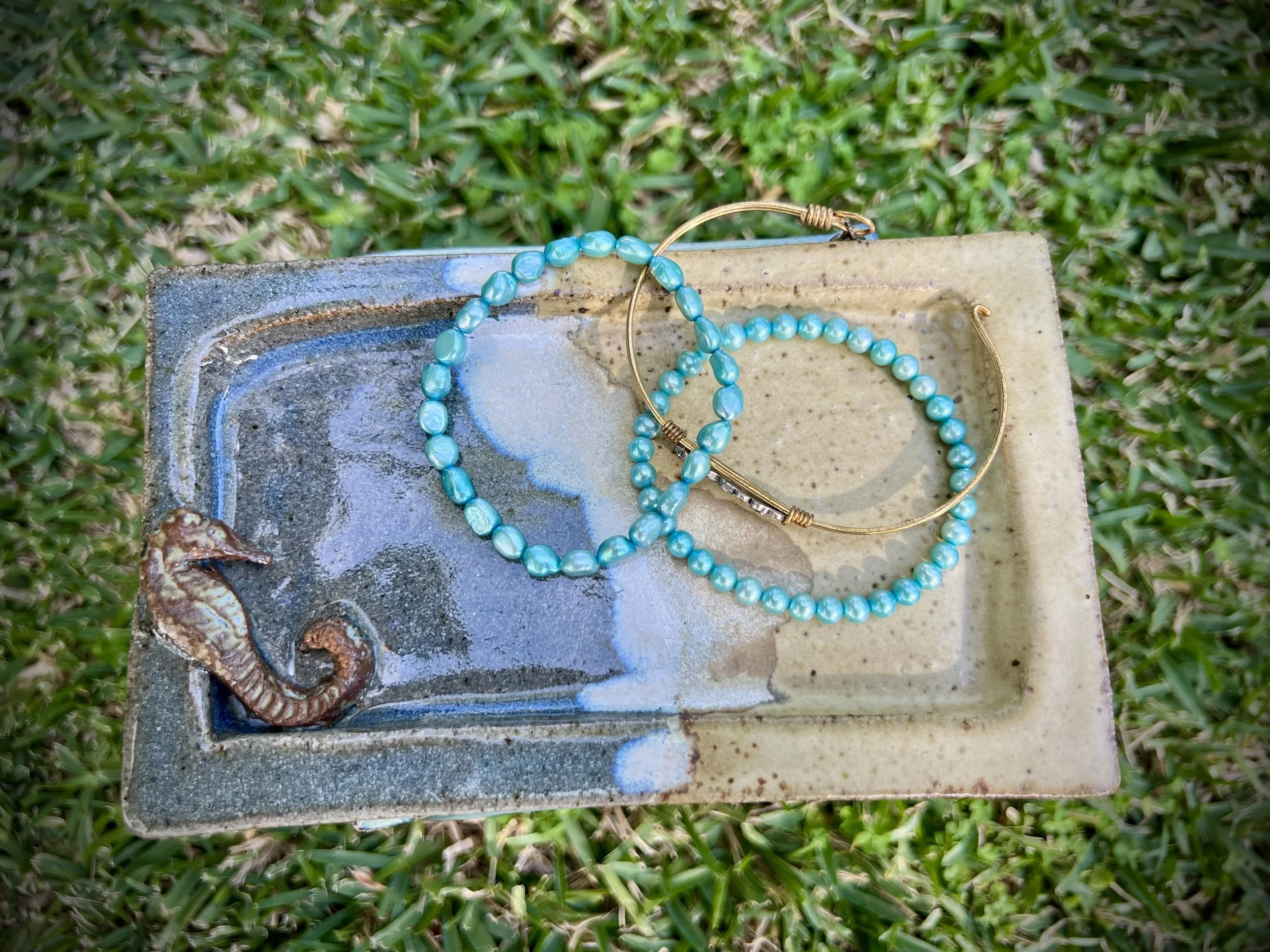 Turquoise beaded bracelets and gold hoop earrings on a decorative ceramic dish with a seahorse design, placed on grass.