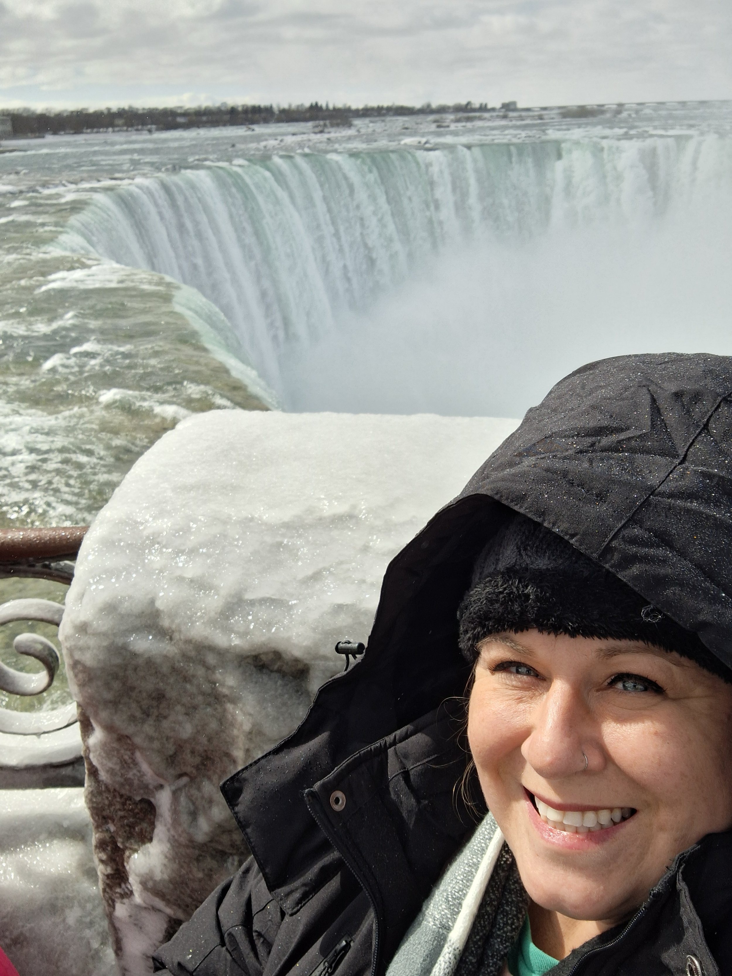 A smiling woman taking a selfie near Niagara Falls with part of a person’s face, snowy rocks, and a cloudy sky in background.