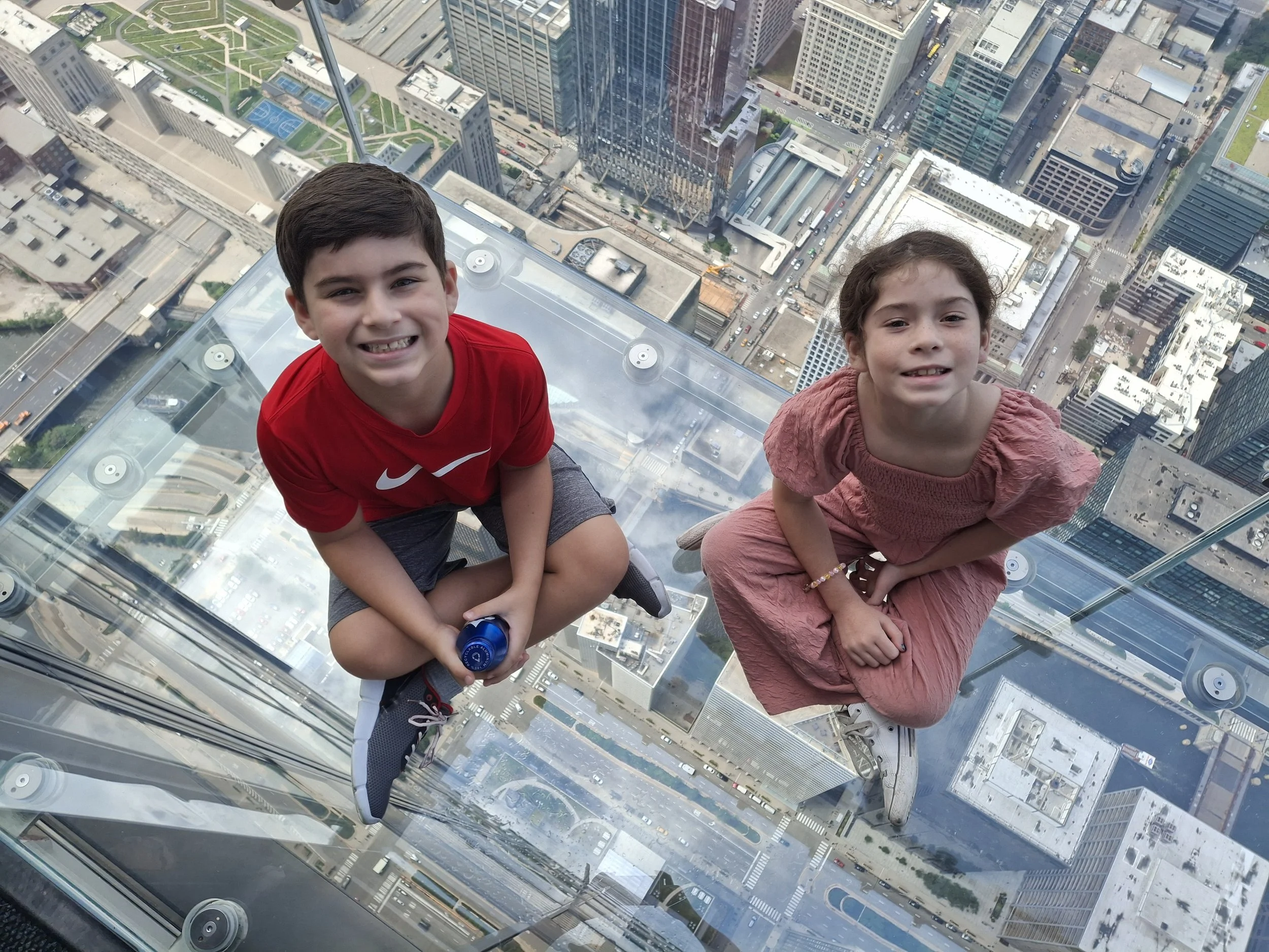 Two children, a boy in a red shirt and a girl in a pink dress, are sitting on a glass observation deck high above a city, with tall buildings, roads, and parks visible below.
