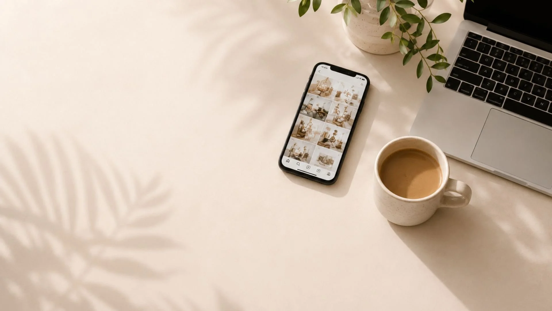 A workspace with a keyboard, a potted plant, a smartphone displaying a grid of interior images, a cup of coffee, and a white surface.