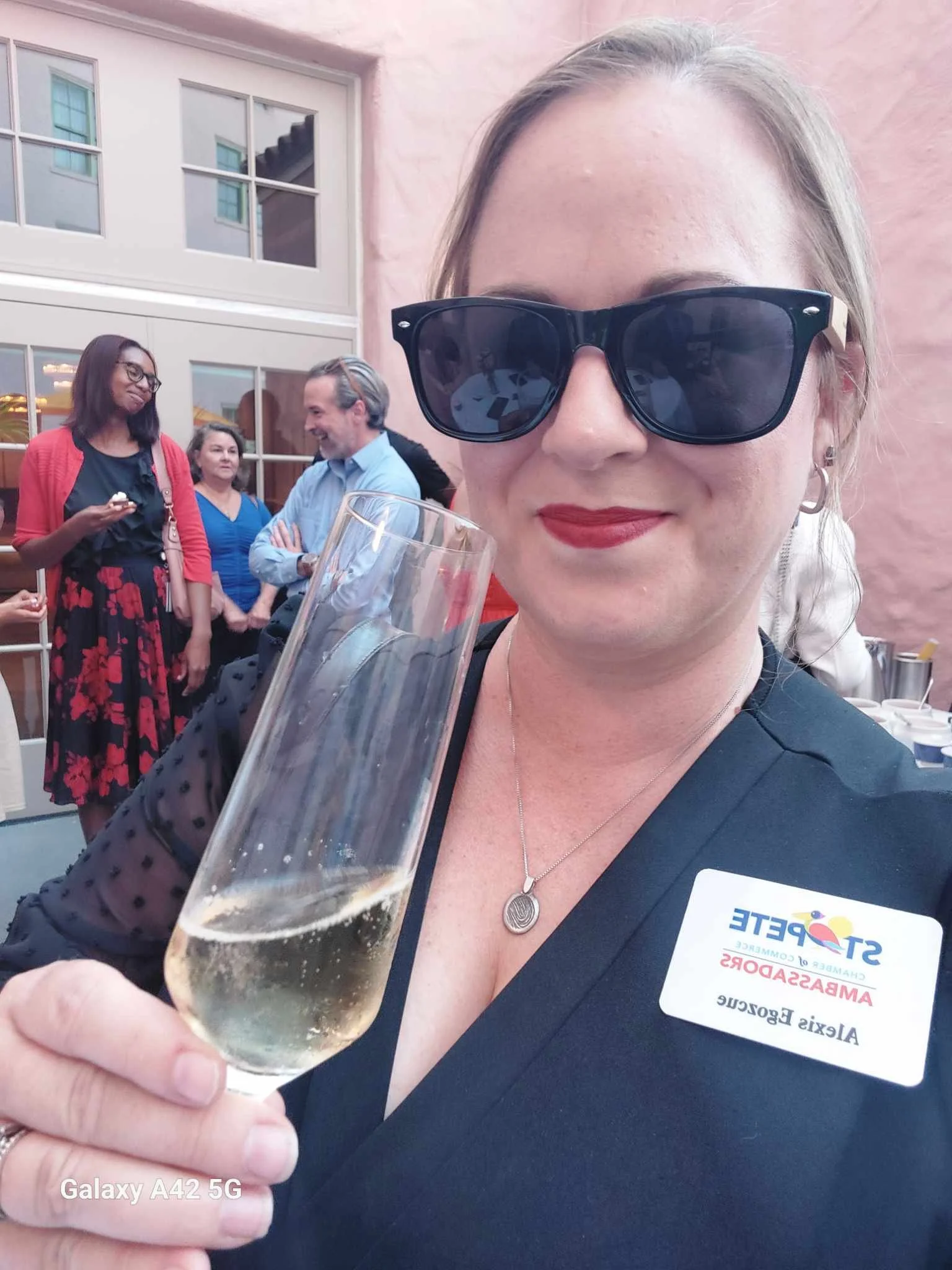 A woman with blonde hair and sunglasses holding a glass of champagne, wearing a nametag with the logo of a company called 'St. Pete' and the name 'Alexis' at an outdoor event with people in the background socializing.
