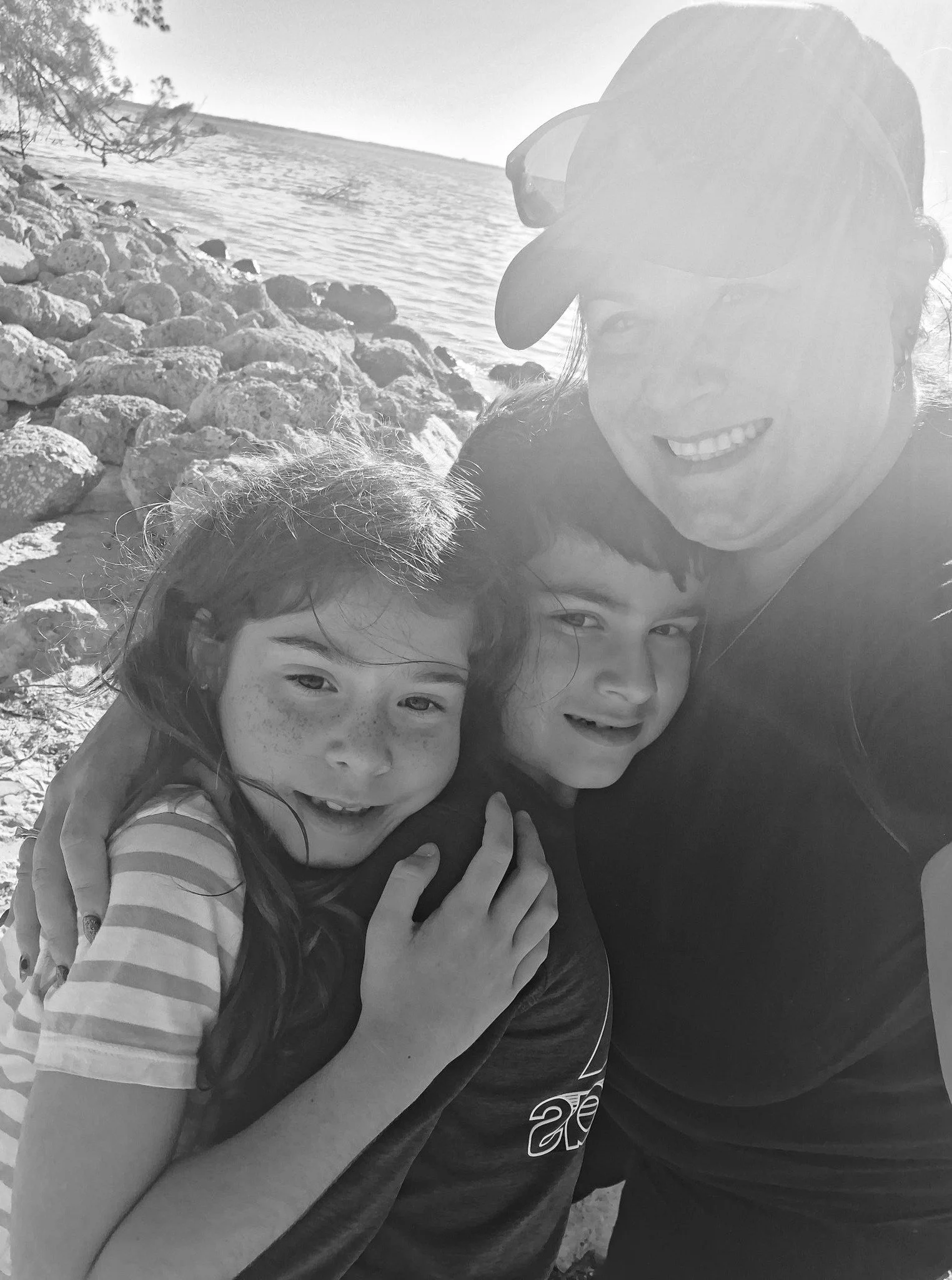 A woman and two children smiling near a body of water, with rocks on the shoreline and a tree in the background.