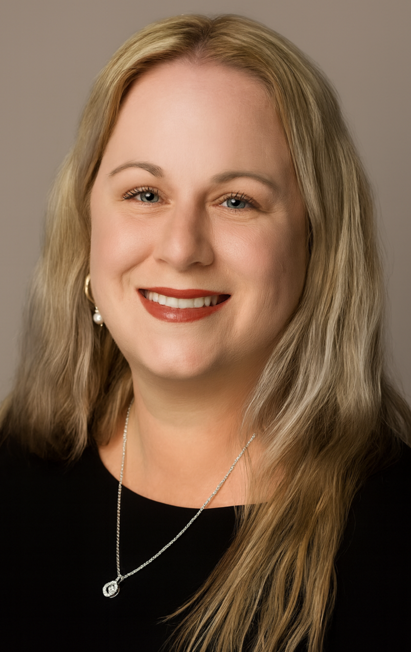 Close-up portrait of a woman with blonde hair, blue eyes, wearing a black top, pearl earrings, and a silver necklace with a round pendant, smiling at the camera.