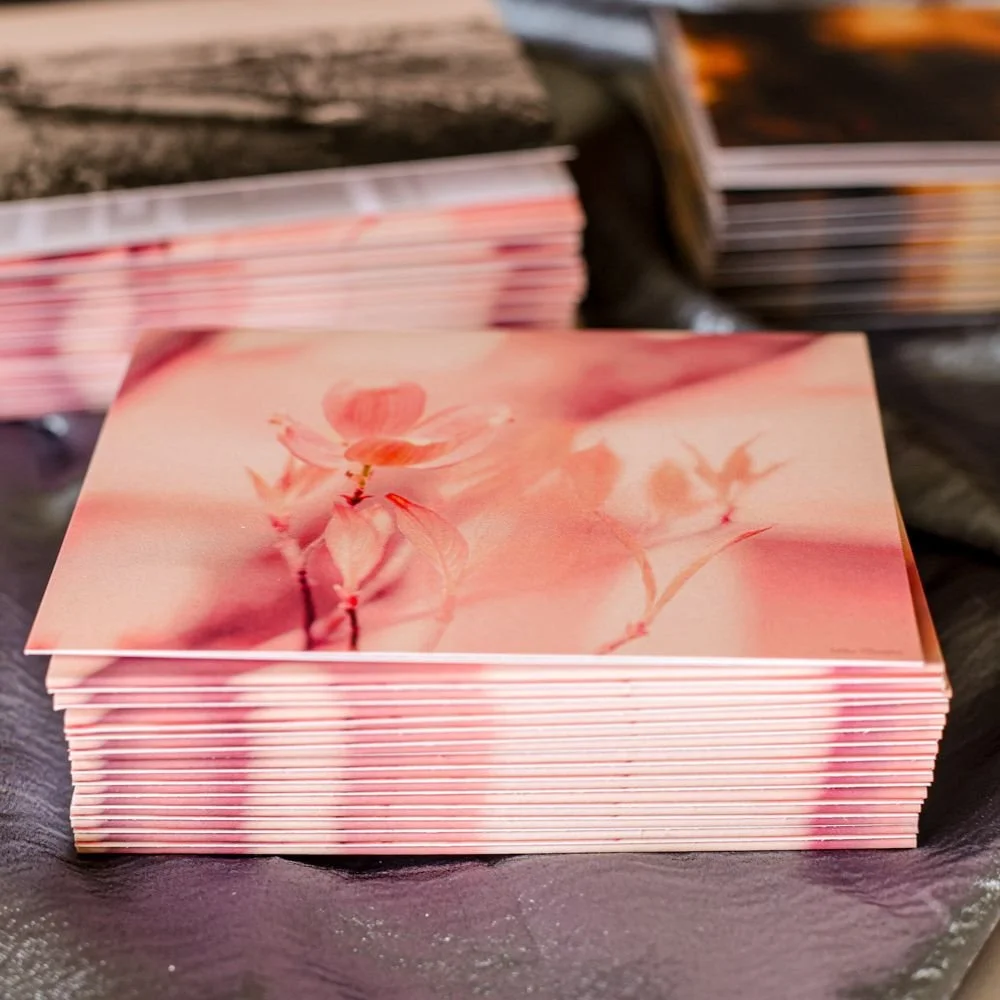 A stack of pink-colored postcards with a soft-focus image of cherry blossoms printed on the top card, placed on a dark surface.