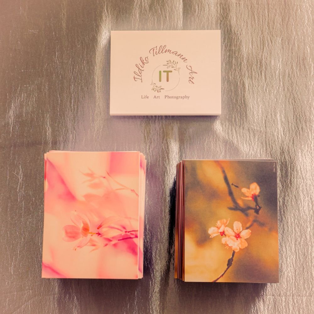 Three stacks of postcards or prints on a metallic surface, with a small box or card above labeled 'Hilary Tillmann Art' including the words 'Life, Art, Photography'.