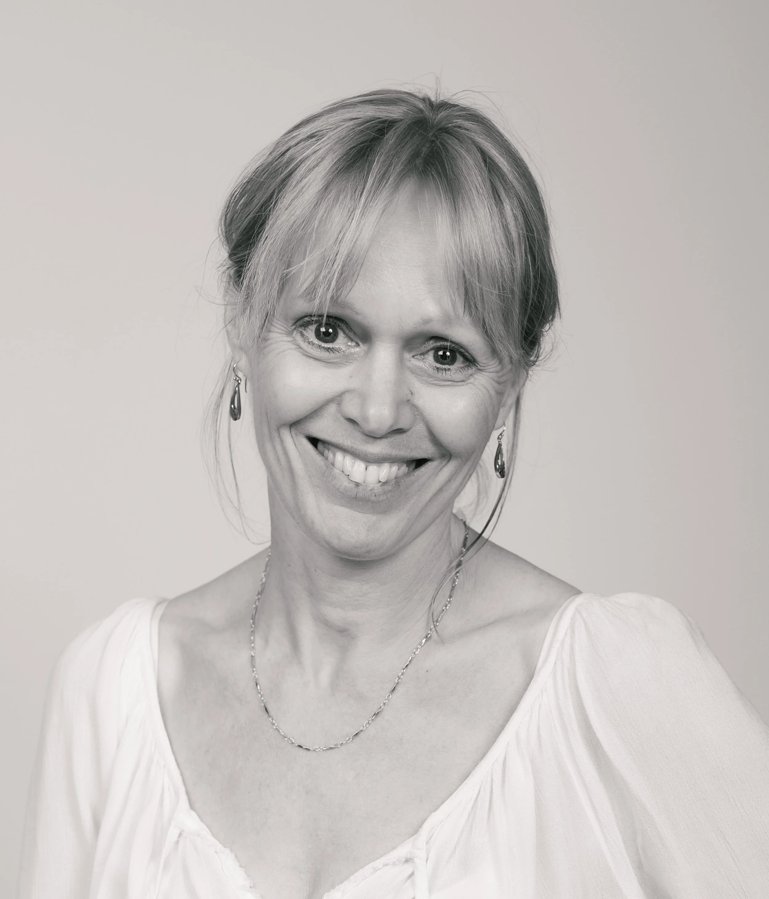 A smiling woman with short, light-colored hair wearing earrings, a necklace, and a light-colored blouse.