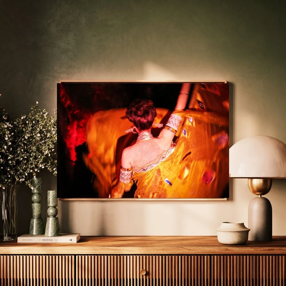 A living room with a large framed art piece of a woman dancing in a yellow dress on the wall. There is a wooden sideboard below the art, decorated with a white book, two vases, a lamp, and a white bowl. A vase with greenery is on the left side of the