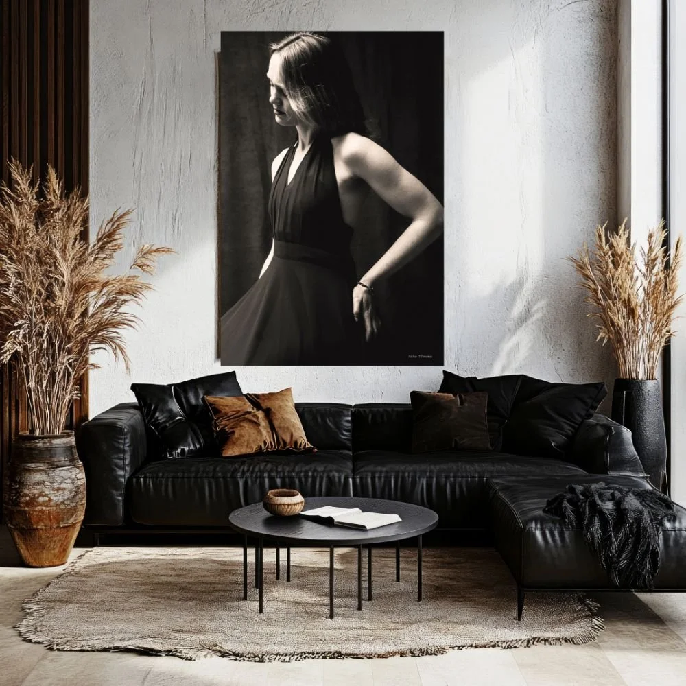 A modern living room with a black leather sectional sofa, black and brown throw pillows, a round black coffee table with a book and a small bowl, large vases with dried plants on each side, and a large black-and-white portrait of a woman hanging on t